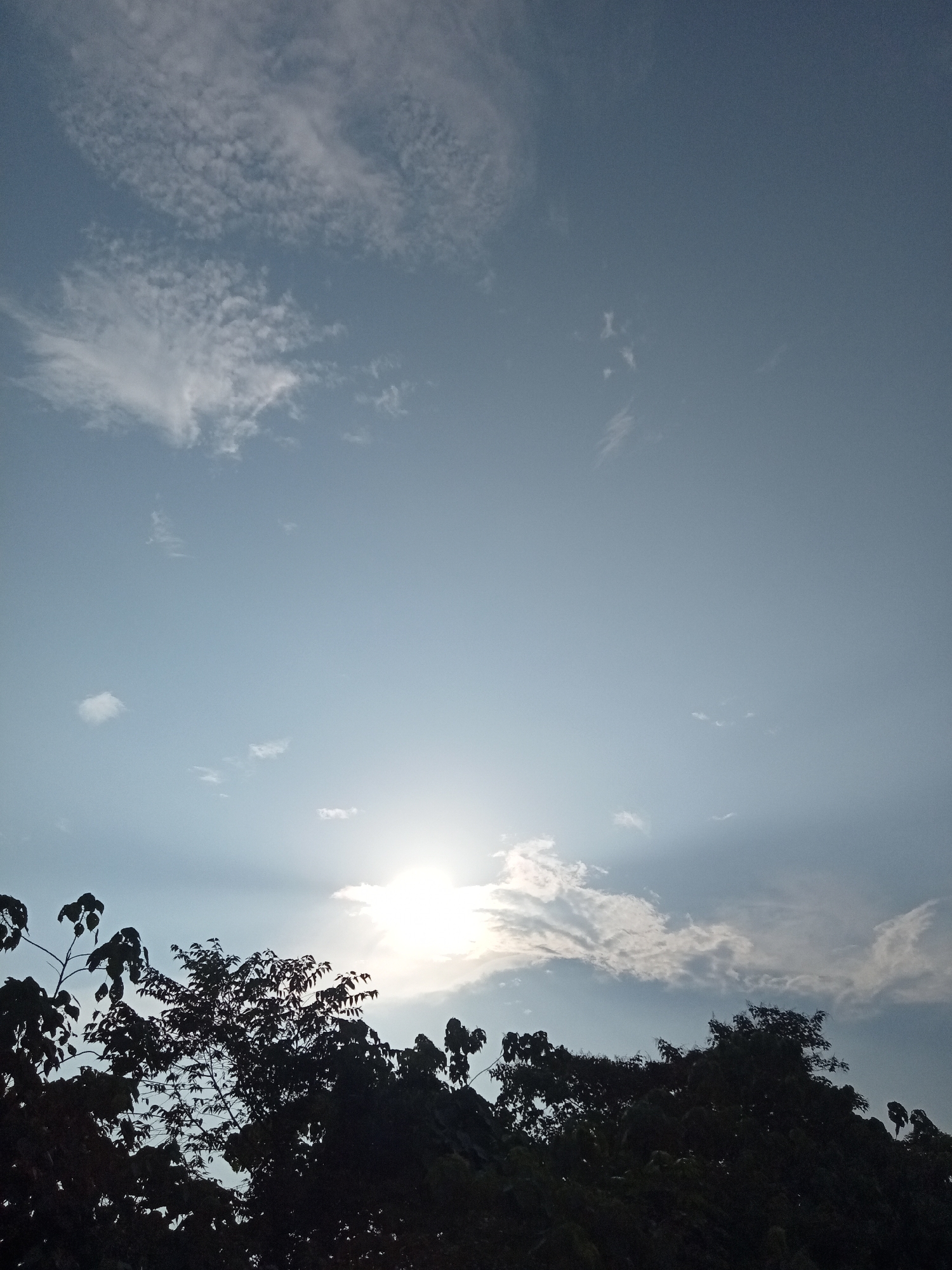 The image captures a clear sky with scattered clouds above a silhouette of trees. The sun is partially obscured by clouds, casting rays of light. The scene appears to be an outdoor environment, possibly a park or a residential area in Teluk Kenidai, Indonesia, given the vegetation and sky. There are no people or animals visible in the image. The lighting suggests it might be late afternoon or early morning.