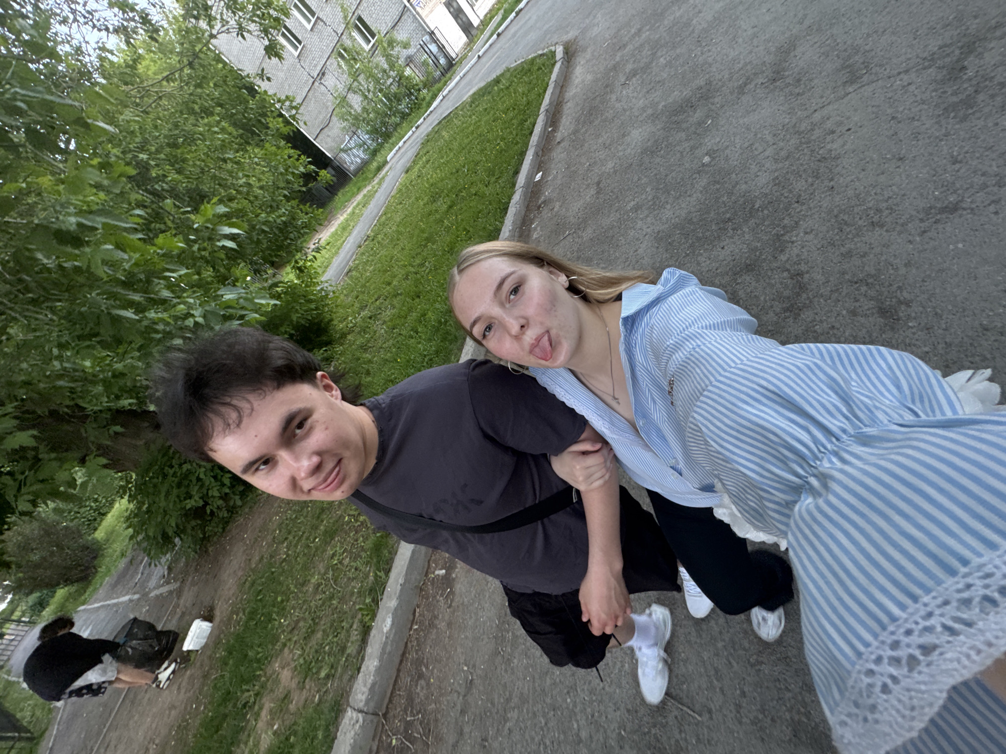 A photo of a young man and woman, shot at an angle in Perm, Russia. The man has dark hair and wears a dark grey T-shirt, black shorts, and white sneakers. He has a black crossbody bag. The woman has blonde hair and wears a striped blue and white blouse, black pants, and white sneakers. She is sticking her tongue out. They stand on a concrete walkway near a grassy area and trees. In the background, there is a building. In the lower left corner of the image, an unidentifiable person is tending to black trash bags.