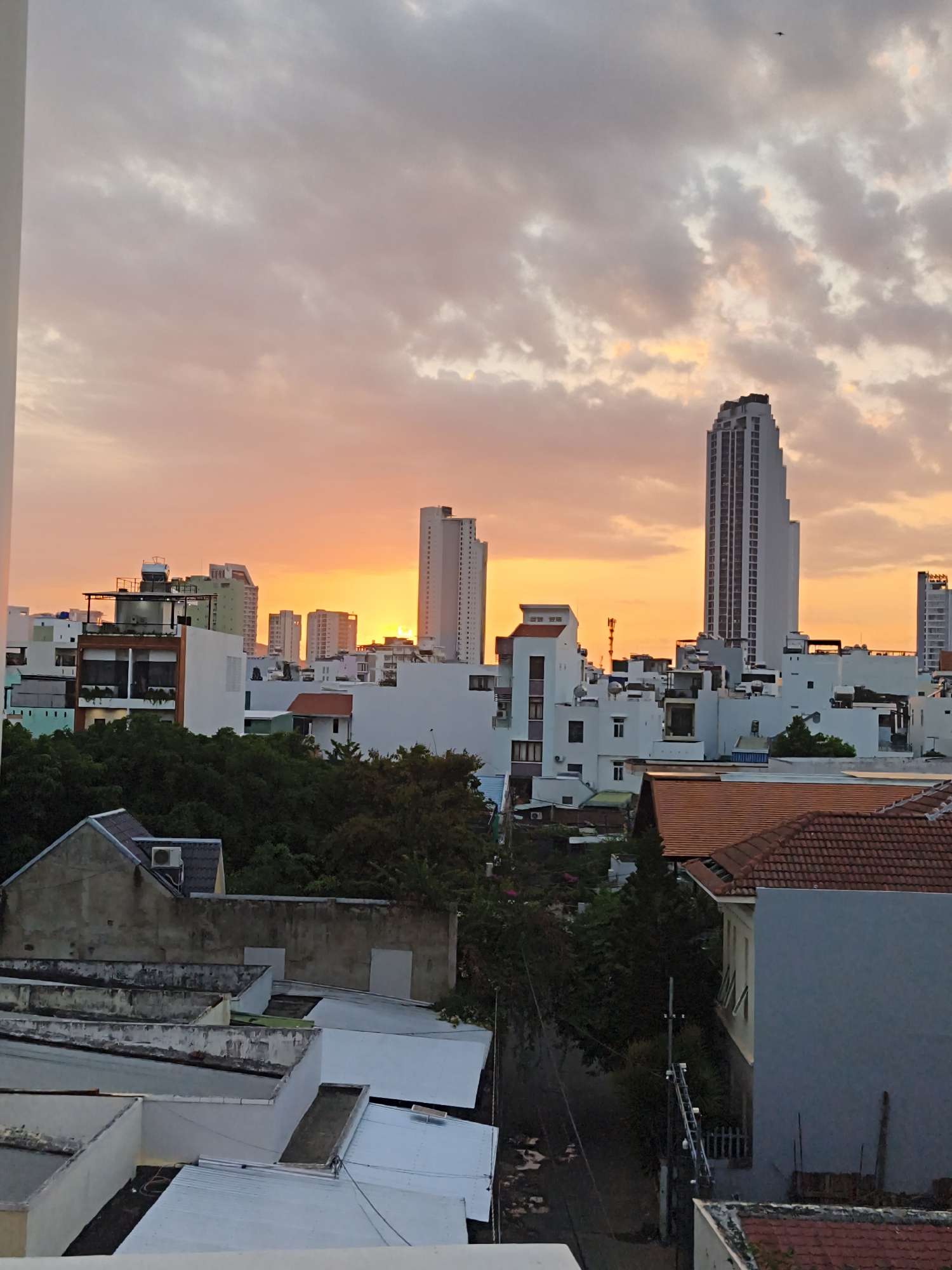 A panoramic cityscape view of Bac Nha Trang Ward, Vietnam, is captured during a sunset. The sky is painted with hues of orange and pink, with scattered clouds. In the midground and background, modern buildings, including several high-rise structures, dominate the skyline. The foreground features a complex of rooftops of various residential buildings, characterized by white and terracotta tiled roofs, some with visible air conditioning units and water tanks. Dense green trees are interspersed between the buildings, adding a touch of nature to the urban environment. The overall scene depicts a bustling urban area at the end of the day, with the warm glow of the sunset illuminating the architecture.