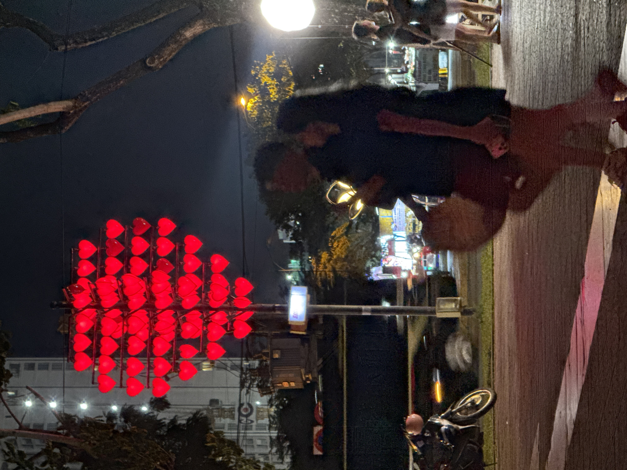 At night in Đà Nẵng, Vietnam, a couple embraces on a sidewalk near a large heart-shaped light display composed of numerous red heart lights. The couple is illuminated by overhead lighting. A street and buildings are visible in the background. A motorcycle is parked nearby. Pedestrians are walking in the background.