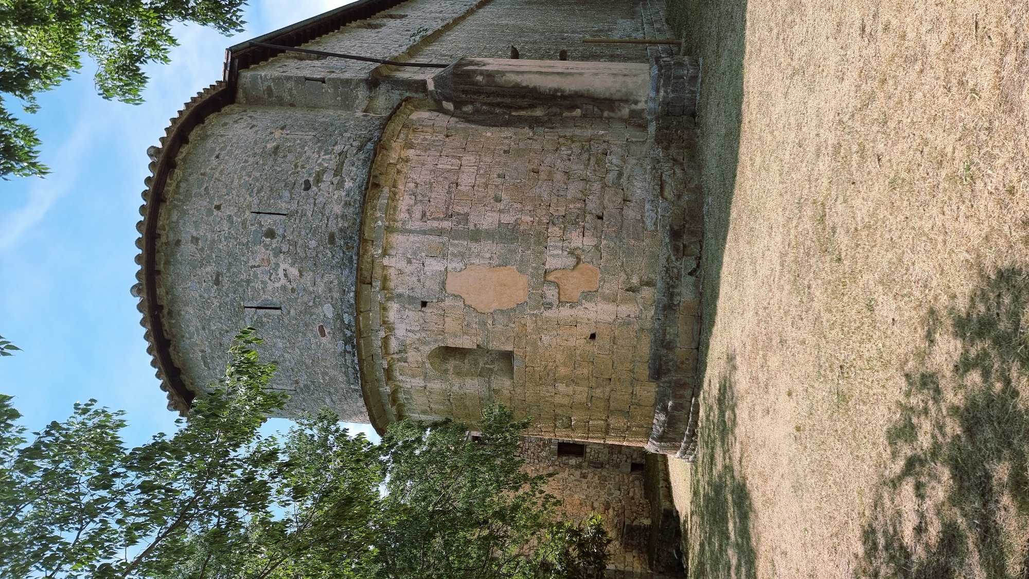 The image shows the apse of a stone church, likely the Church of Sant Mateu, located in Sales de Llierca, Spain. The apse is circular and constructed from large, rough-hewn stones. It has a tiled roof. Several small, narrow openings are visible on the upper part of the apse. There is a small, arched window higher up on the curved wall, and some areas of exposed plaster are visible on the stone. The base of the apse is built on a lower stone foundation. To the left and behind the apse, another stone building with a window is partially visible. The church is surrounded by dry, golden grass, with dappled shadows from trees cast across the foreground. Green foliage from trees frames the top of the image. The sky is blue with some scattered white clouds. The scene is static and depicts an exterior view of the historical church structure.