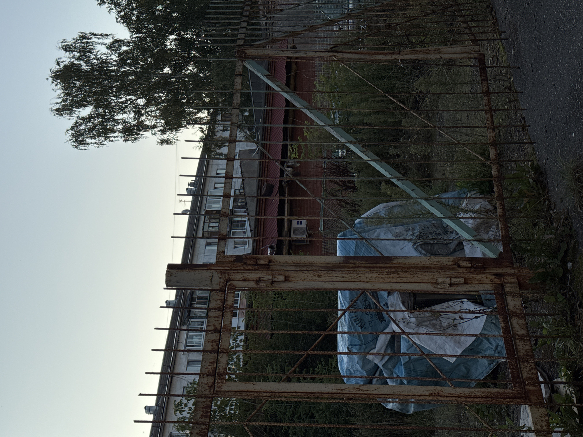 The image shows a rusty metal gate in front of a multi-story building with a red-tiled roof. Behind the gate, there are several large objects wrapped in blue tarpaulin, suggesting they are stored materials. The building appears to be residential, with multiple windows visible. Trees and overgrown vegetation are also present in the scene, creating a slightly unkempt environment. The sky is overcast, indicating a possibly cloudy day. The overall impression is one of an industrial or utilitarian area, possibly on the outskirts of an urban setting. The location is identified as Izhevsk, Russia.