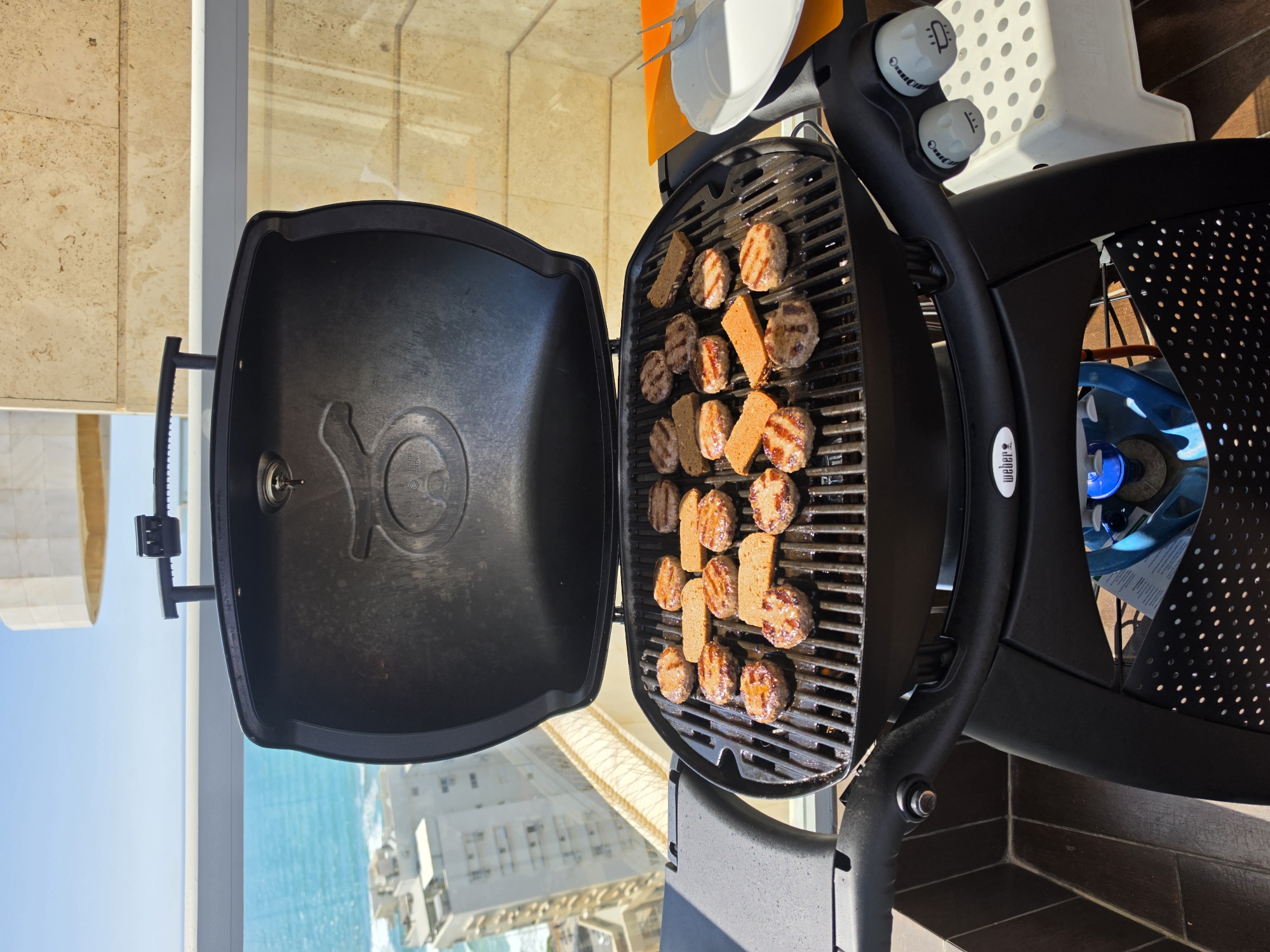 The image shows a portable Weber gas grill positioned on what appears to be a balcony overlooking the city of Bat Yam, Israel. The grill is open and contains an assortment of small, grilled meatballs and pieces of bread. The background features a cityscape with the Mediterranean Sea visible in the distance. The grill is equipped with two control knobs and is adjacent to a white, perforated plastic chair. A propane tank is situated in the grill's frame. A plate with utensils is positioned atop the grill. The surrounding structure has light-colored stone or tile cladding.