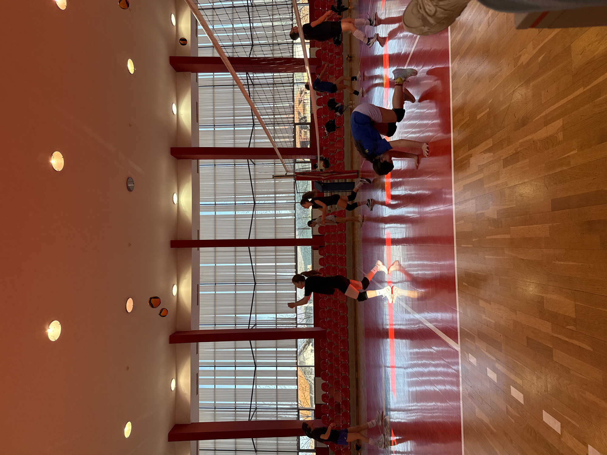 The image shows a volleyball court in what appears to be an indoor gymnasium in Etropole, Bulgaria. Several volleyball players are engaged in stretching and diving exercises. The gymnasium has red floors and red support beams. Large windows run along one side of the gymnasium. There are rows of red seats visible. The lighting is provided by numerous circular fixtures on the ceiling and walls. A volleyball net is strung across the center of the court. The image is slightly rotated.