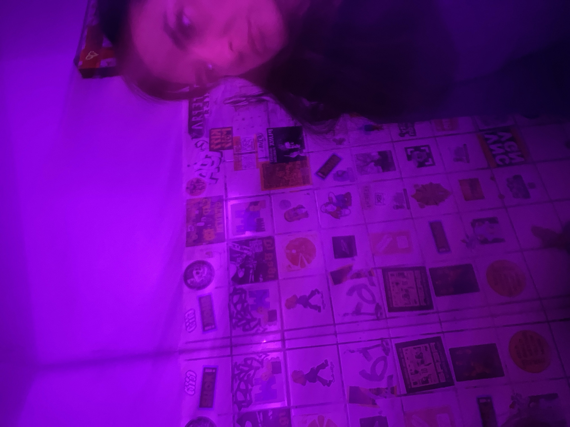 A blurred image depicts a person partially visible at the top of the frame, with their head angled downwards. Below them is a wall covered with square tiles, each bearing various stickers or images. The overall lighting is predominantly purple. It is not possible to determine the location or further details from this image.