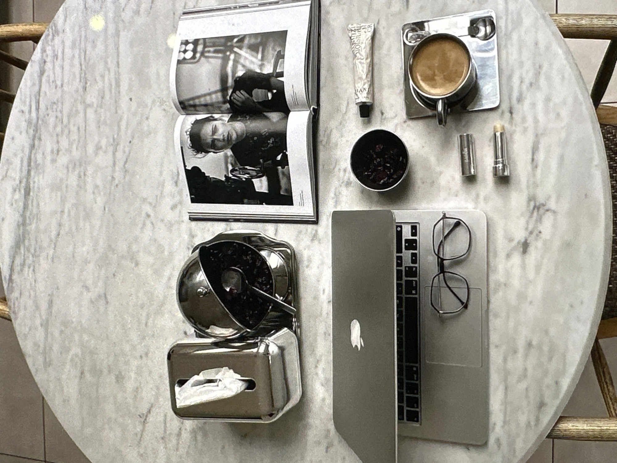 The image depicts an overhead shot of a round marble table with various items scattered across it. A book, opened to a black and white photograph of a person, lies at the top left. Next to it are a tube of lotion, a small container of dark, possibly dried flowers or tea leaves, a cup of coffee on a square metal coaster, and two lip balms. Below these items are a silver candy dish with candies and a tissue box. A partially open Macbook laptop sits next to the candy dish, with a pair of glasses resting on it. The table appears to be situated in a café, with parts of a wicker chair visible at the edges of the frame. There is no indication of this location being in Yerevan, Armenia.