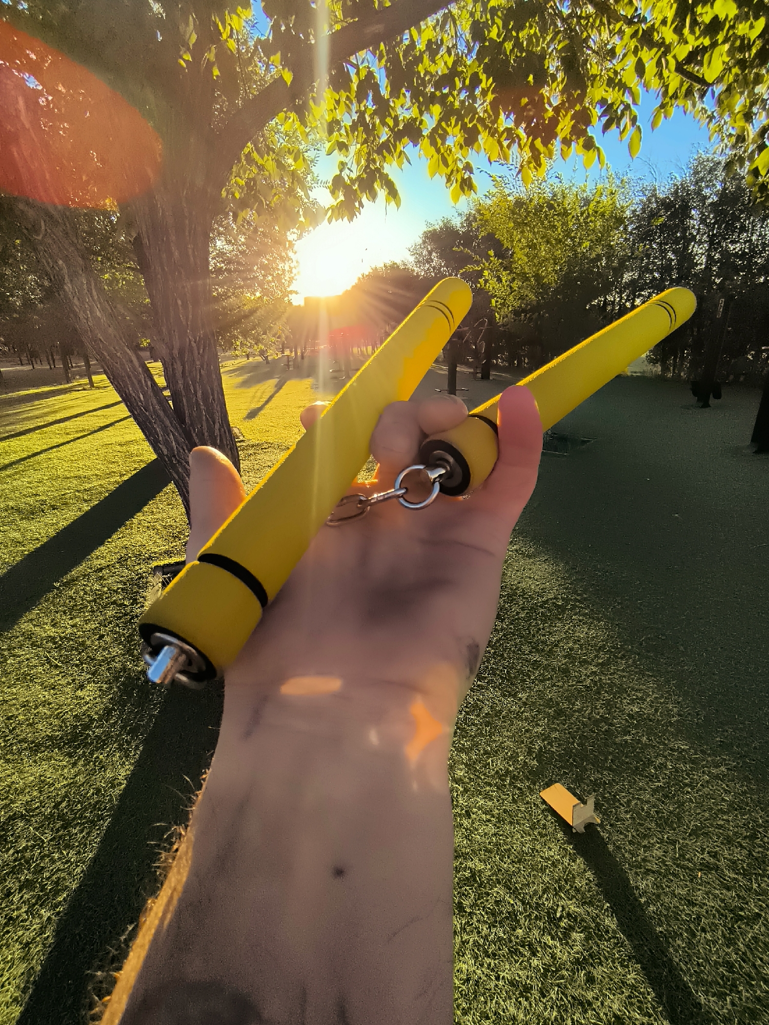 A first-person perspective shot captures a hand holding a pair of bright yellow nunchucks in a park-like setting. The nunchucks have black accents near the ends, which are capped with silver metal. A metal ring connects the two segments of the nunchucks. The hand holding the nunchucks appears to have some smudges on it. The background shows a well-maintained grassy area with shadows cast by trees. Sunlight streams through the leaves of a large tree in the foreground, creating lens flares and a bright, warm glow. In the distance, more trees and a clear blue sky are visible. There are no discernible landmarks that would indicate the specific location of Qarağandı city administration, Kazakhstan.