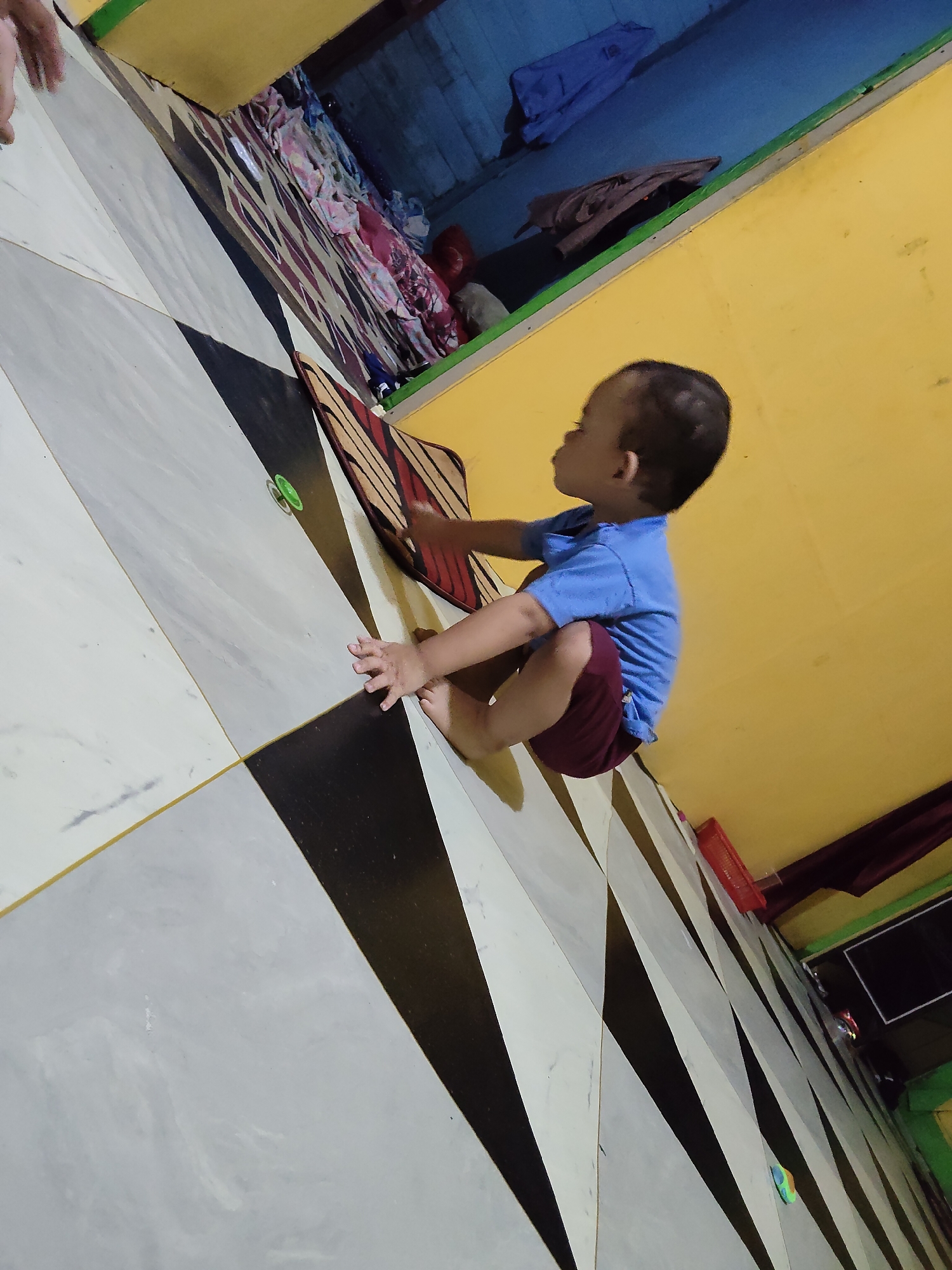 A toddler is sitting on a floor with a geometric tile pattern. The toddler is wearing a blue t-shirt and maroon shorts and is facing to the left. The floor is made up of alternating black and white diamond shapes, with thin yellow grout lines. In the background, there is a yellow wall and a blue wall, with some folded clothes and other items on a shelf. There is also a red basket on a shelf. The image appears to be taken indoors. The location is in the city of Tanah Grogot, Indonesia.