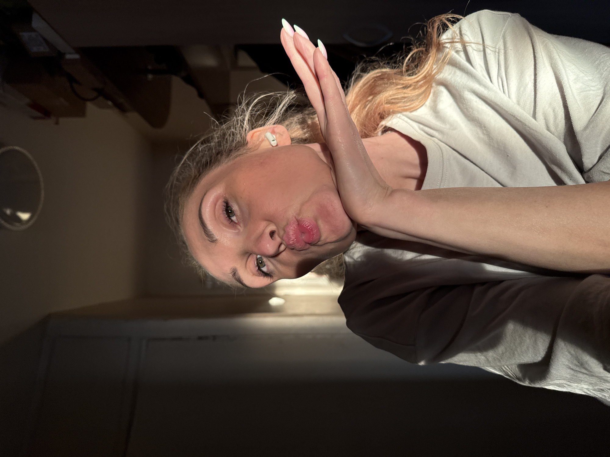 A young woman is captured in a close-up photo, rotated 90 degrees counter-clockwise, likely taken indoors in Chaikovsky, Russia. She is making a playful "pouty" or "kissing" expression towards the camera, with her lips puckered. Her green eyes are open and engaging. Her light-colored hair, possibly blonde or light brown, is tousled around her face and shoulders. An earbud is visible in her left ear. Her right hand is raised, with manicured nails featuring light tips, resting against the side of her face. She is wearing a light-colored, possibly white or light gray, t-shirt that shows soft folds.
The lighting is dramatic, casting strong highlights on her face, hair, and clothing, while the background is mostly dark and indistinct, creating a high-contrast effect. Faint outlines of furniture or room details are barely visible in the darkened background, including a circular object. The image focuses almost entirely on the woman, emphasizing her expression and pose. No text is visible.