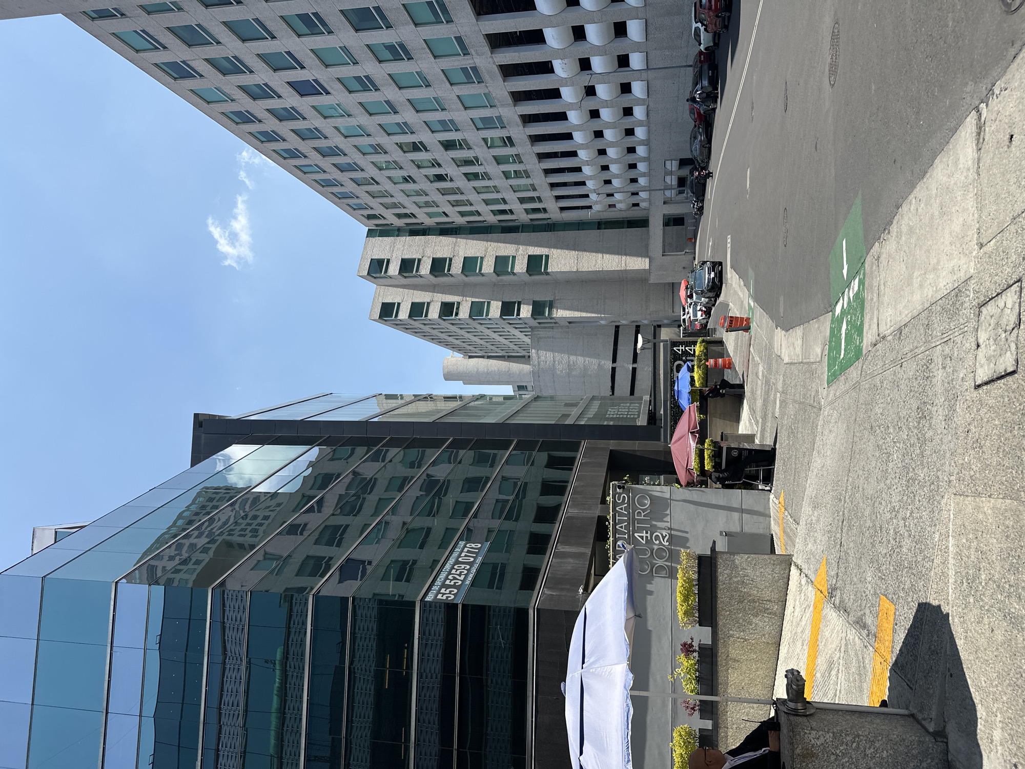 A street scene in Mexico City, Mexico shows modern office buildings, including a glass facade and a concrete building with modular windows. The street has a green arrow marking a lane, parked cars, traffic cones, and cafe umbrellas. A sign on one building reads "ARIATAS CUATRO DO". There are some people sitting or standing near the storefronts. The sky is clear blue with a few clouds.