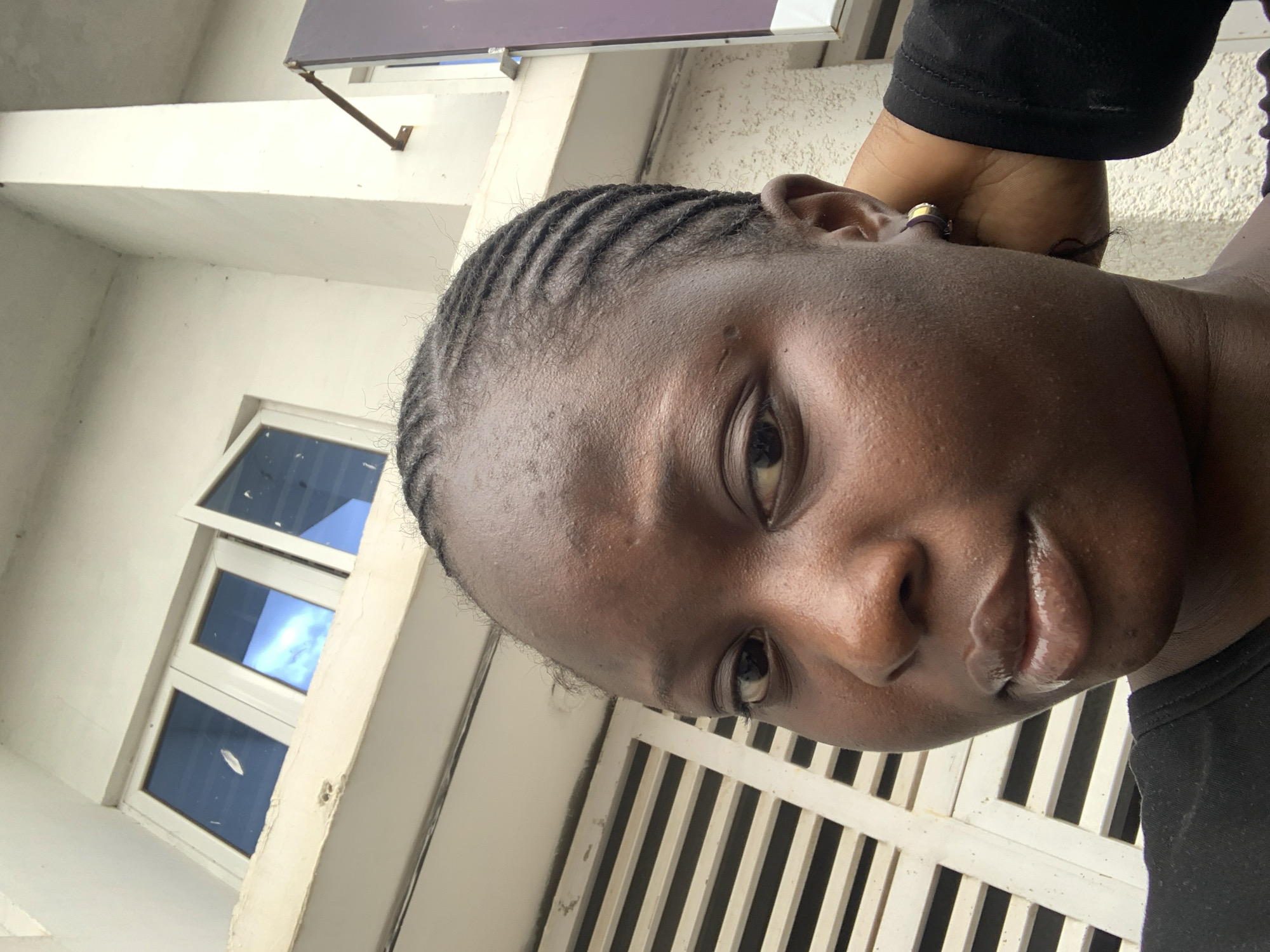 A close-up, selfie-style shot features a young Black woman with braided hair and a black shirt, positioned outdoors in front of a building with windows and white shutters. The woman's face fills most of the frame, with her right hand supporting her head. The background includes a white building facade with reflective blue windows and horizontal white slats. The lighting suggests it is daytime.