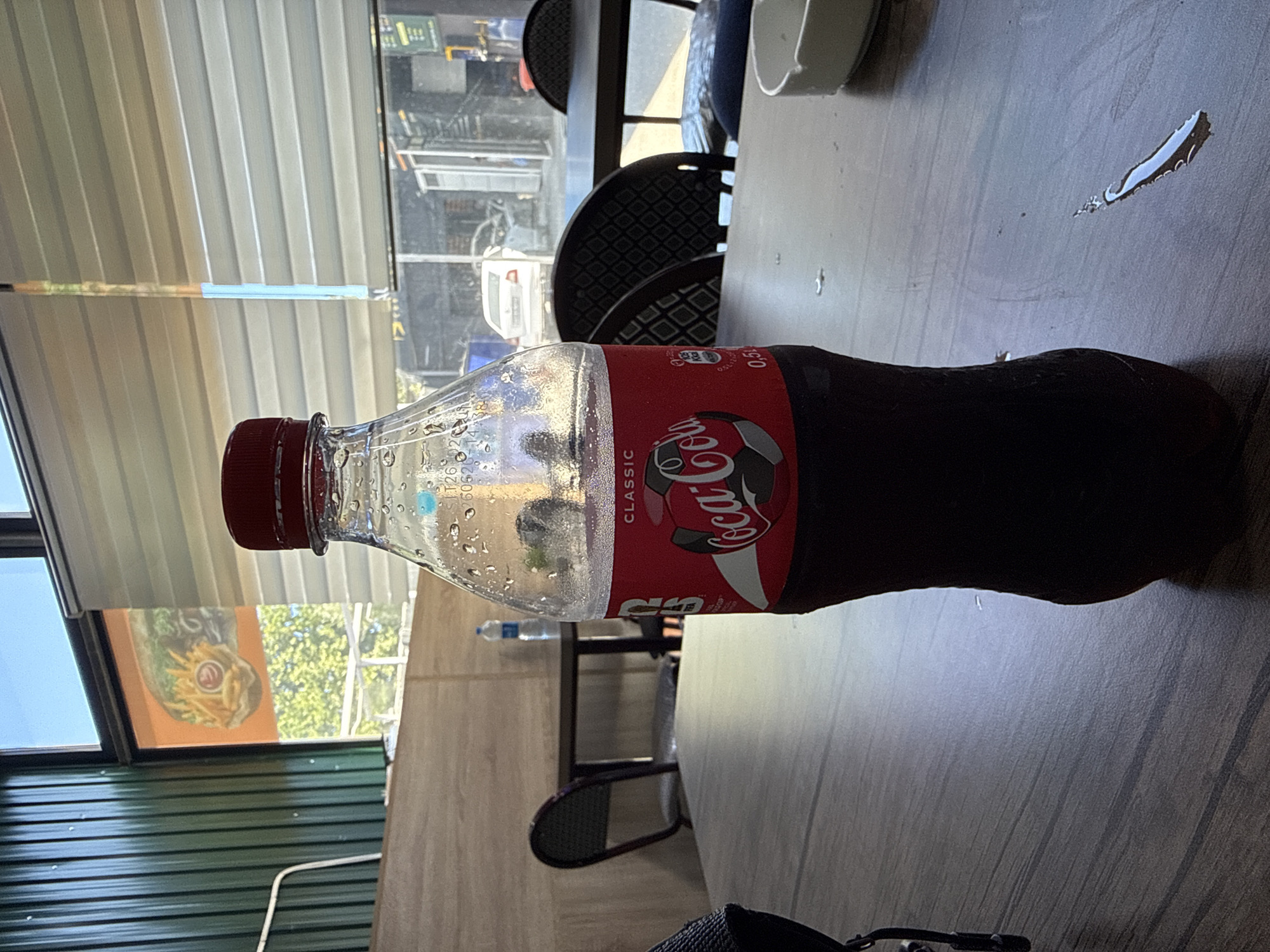 A close-up shot focuses on a plastic bottle of Coca-Cola with a red cap, condensation visible on its surface, positioned on a wooden table. The bottle is centrally located and fills a significant portion of the frame. The table surface exhibits a wood grain pattern and appears to have some spilled liquid. In the background, through a window with blinds, a glimpse of an outdoor scene is visible, including trees and buildings, suggesting an urban or suburban setting. The overall impression is that of a casual dining or resting environment. The location is identified as Khatay Raion, Azerbaijan.
