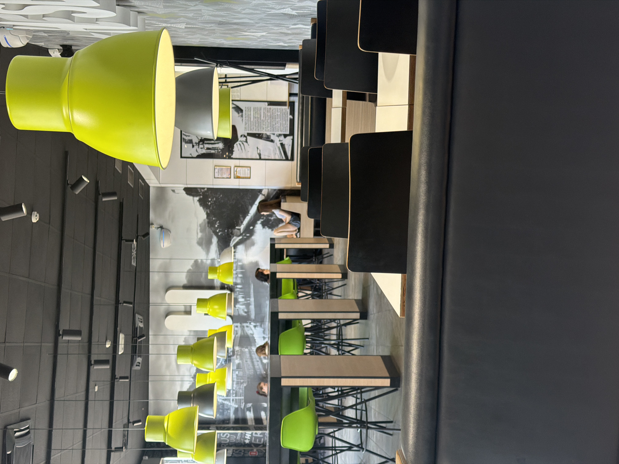 A low-angle shot shows a row of tables and chairs in a modern restaurant. The tables are light brown with black trim, and the chairs are bright green. The tables are set up in a long, communal style, with chairs on either side. In the background, there are large black lamps hanging from the ceiling, and a mural depicting a black and white cityscape. A few people are visible sitting at the tables, and some are walking in the background. The restaurant appears to be well-lit and modern. The image is oriented vertically. No visible text is legible.