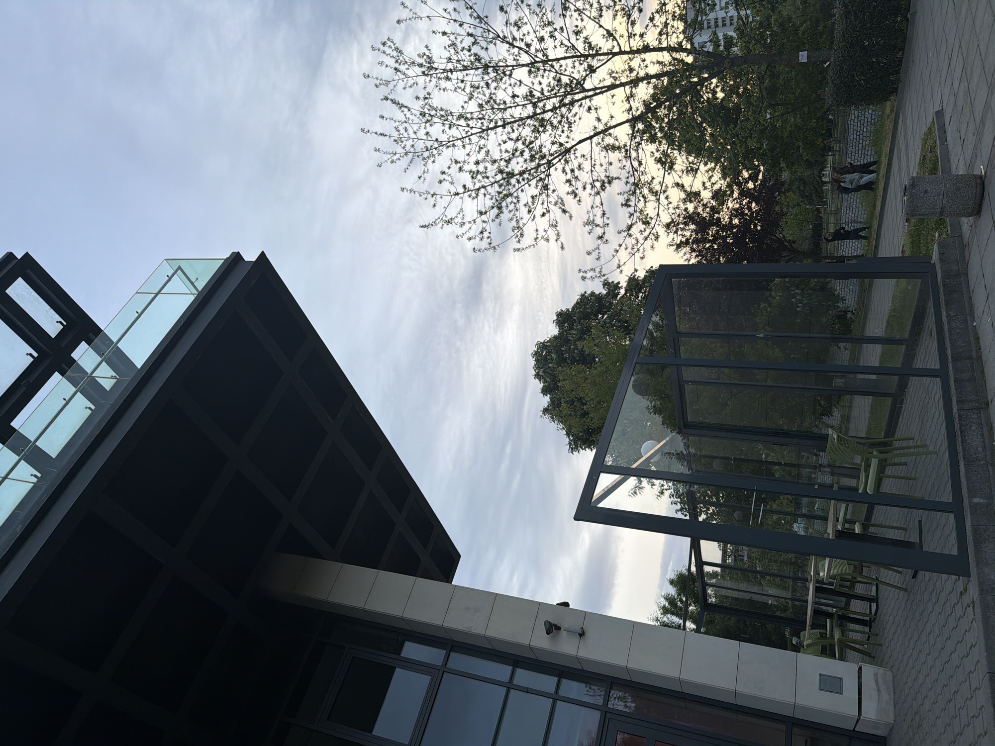 The image captures a low-angle view in an urban setting, likely Blagoevgrad, Bulgaria, given the user's specification. Dominating the left side is a modern building with a dark, geometric facade and large glass panels. A security camera is visible on the building's exterior. To the right, a glass-enclosed structure, possibly a covered patio or bus stop, is situated on a paved area. The sky is overcast, with streaks of clouds. In the background, trees are visible, and what appears to be a group of people are in the distance near the greenery. The image is angled, giving a dynamic perspective of the scene.