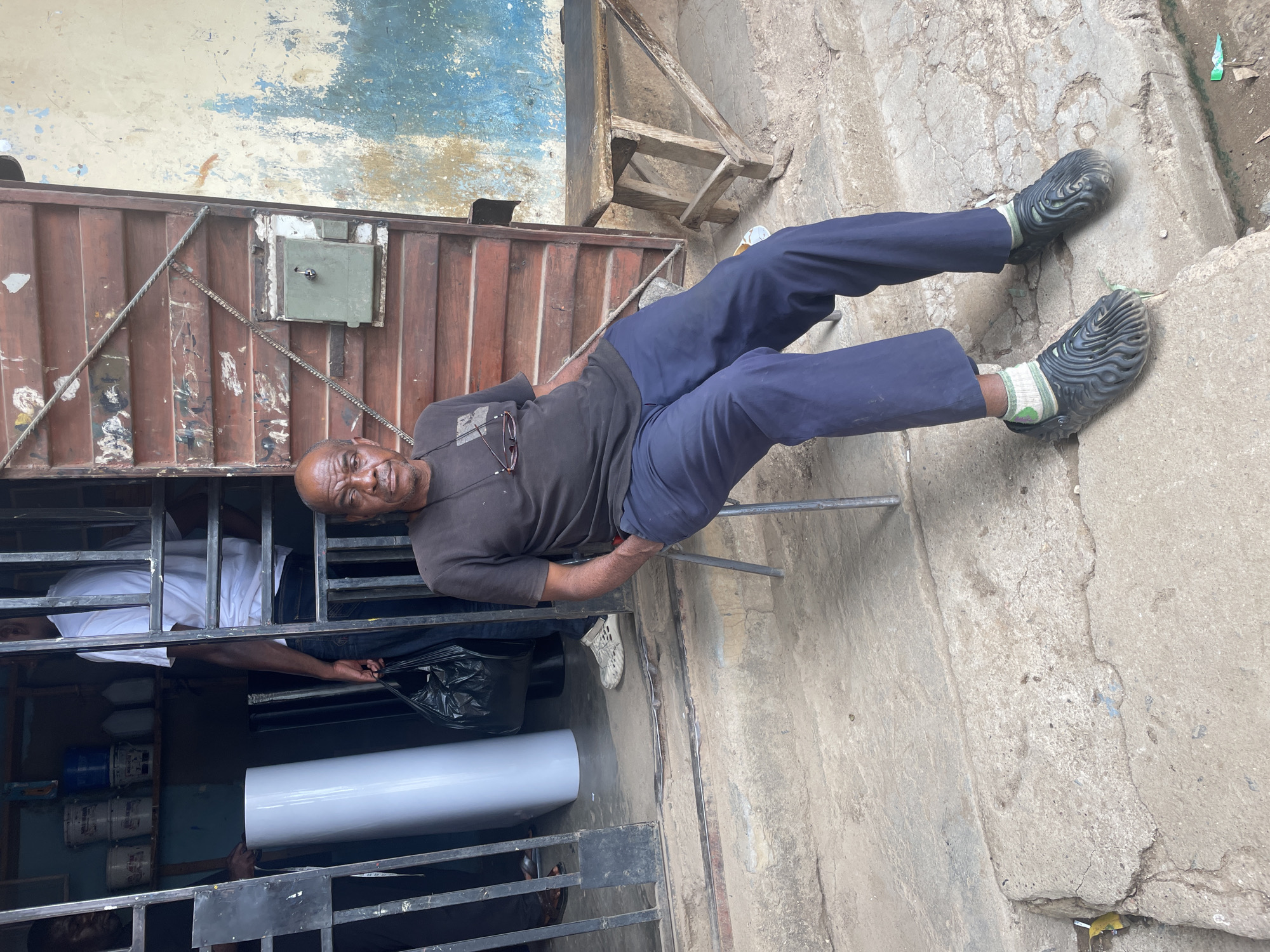 The image depicts a man sitting on a metal chair, outdoors, on a concrete surface. He is wearing dark blue pants, a dark t-shirt, and dark-colored shoes with patterned soles and green socks. A pair of sunglasses are visible hanging on his shirt. The background shows a weathered building with a rusted, brown, corrugated metal gate. Through the gate, other people can be seen inside a building, including one person in a white shirt and another holding a long, white cylindrical object. There are paint cans visible as well. The setting appears to be a commercial or industrial area in the city of Jos, Nigeria. A dilapidated wooden stepladder is visible against the wall behind the man.