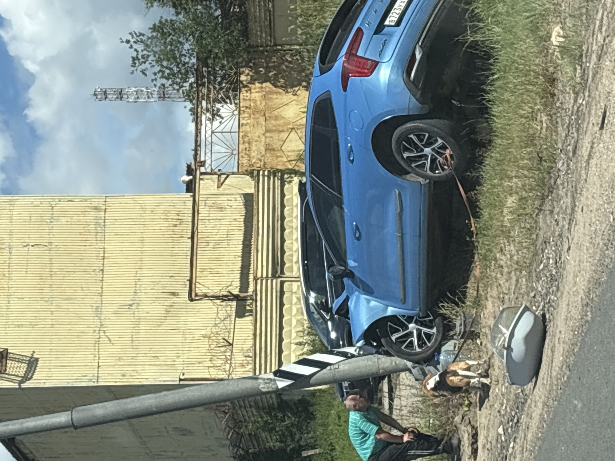 A blue station wagon has crashed into a utility pole on the side of a road. The car's front end is severely damaged, and it is tilted on its side. A man in a green t-shirt and dark pants is leaning against the utility pole, looking at the car. A beagle is standing next to the man, on a leash. A light blue bucket lies on the ground near the car. In the background, there is a yellow corrugated metal building and a tall cell tower. The scene appears to be in an industrial area. The license plate of the car reads B723YX. The location is identified as Novoye, Russia.