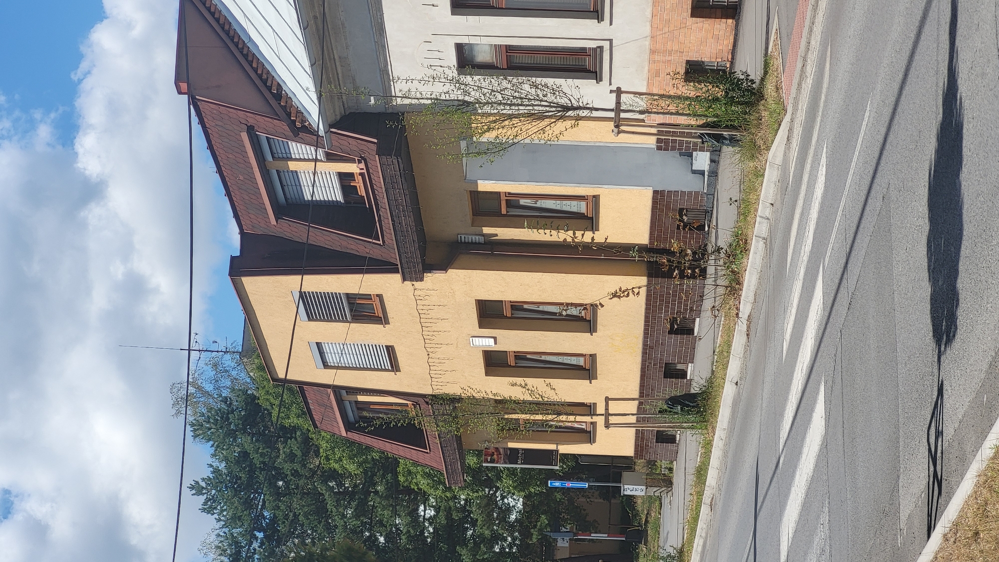 The image captures a street view in Prešov, Slovakia, featuring a two-story yellow building with a mansard roof and dark red shingles. The building has multiple windows with light-colored blinds. Adjacent to it is a lighter-colored building with a similar mansard roof. In the foreground, a pedestrian crossing is visible on an asphalt road. Trees are planted along the sidewalk in front of the buildings, and a traffic sign is present on the left side. The sky is partly cloudy with a bright blue background. The scene appears to be a residential or commercial area on a sunny day.
