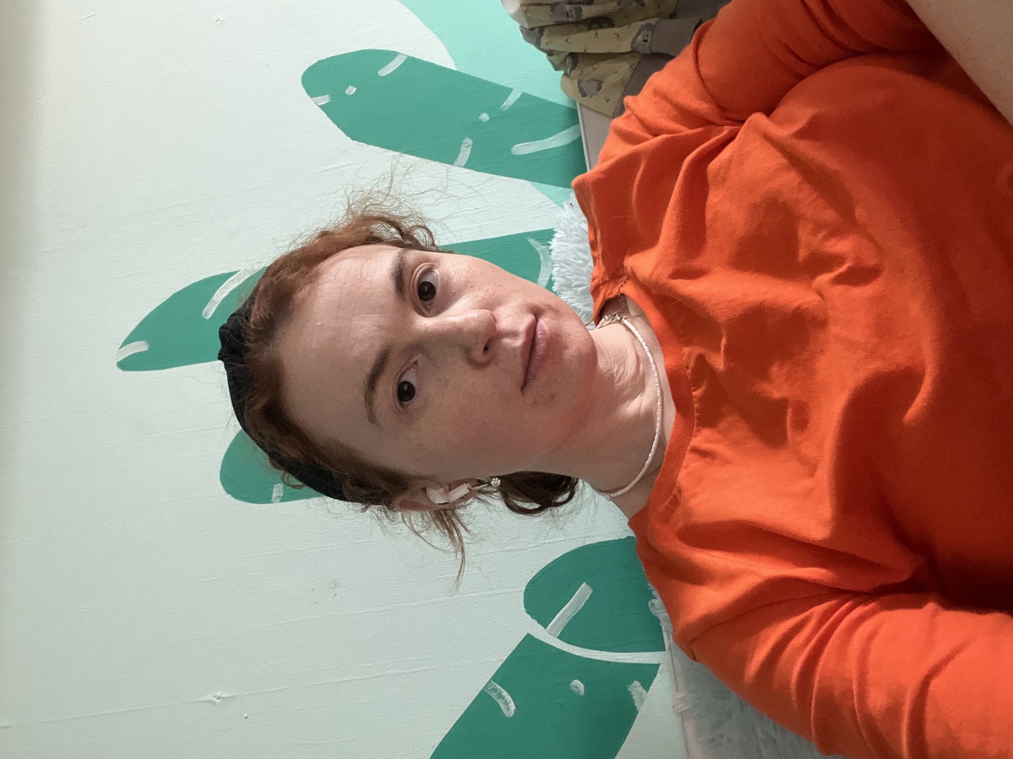 The image displays a person, likely a woman, in an indoor setting. The shot is angled, suggesting a selfie or a photo taken while the individual is reclining. The subject has fair skin, reddish-brown curly hair, and dark eyes, looking directly at the camera with a neutral expression. She is wearing a bright orange long-sleeved shirt, a black headband, and a thin white beaded necklace. Small white earbuds or earrings are visible in her ears.
The background features a light mint green wall decorated with stylized, darker green shapes that resemble large leaves or abstract plant motifs, accented with white lines. These patterns suggest a deliberately decorated space, possibly a bedroom or living area. In the upper right corner, a glimpse of light-colored, soft fabric, potentially a blanket or pillow, is visible, further indicating an indoor, comfortable environment. There is no visible text in the image. The overall scene conveys a casual moment within a decorated room in Chaikovsky, Russia.