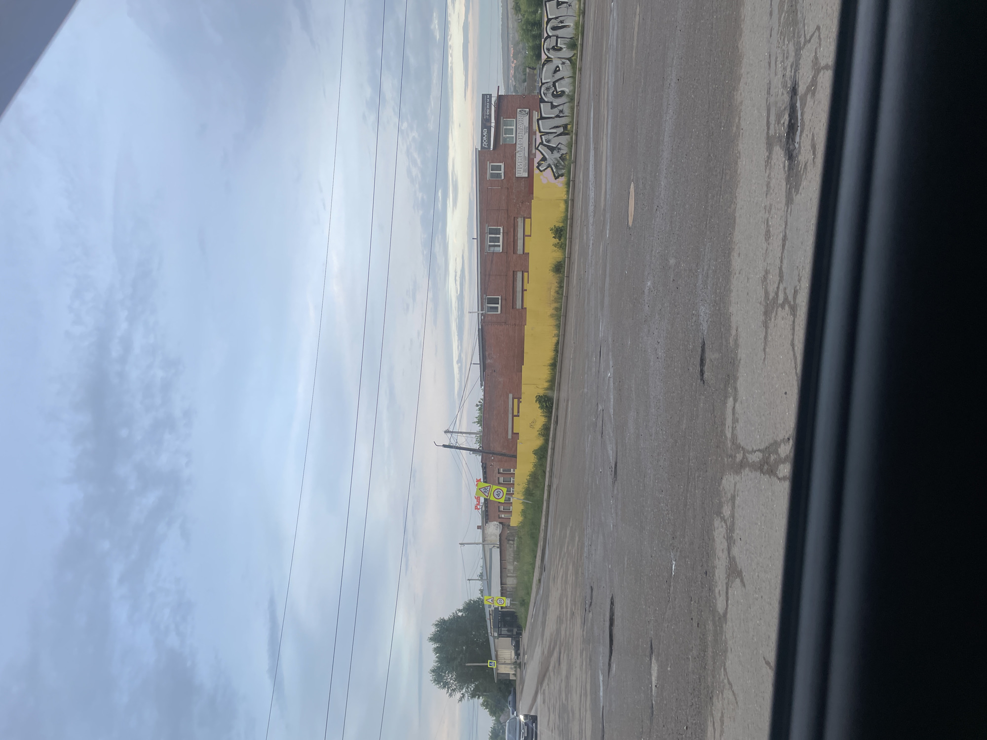 The image depicts an urban street scene in Votkinsk, Russia. A road with visible potholes occupies the foreground, with a black car on the left edge moving away from the camera. The middle ground features a long, brick building with a bright yellow fence along its front. Several yellow road signs with cautionary symbols are visible, one indicating a speed limit of 40. The building has multiple windows and signage, including "ДОМА" (Doma) and "ШИНОМОНТАЖ" (Shinomontazh), suggesting residential and tire service businesses. Graffiti in large white letters is spray-painted on the yellow fence. The background shows a cloudy sky and a distant, hilly landscape. The scene appears to be in an industrial or commercial area on an overcast day.
