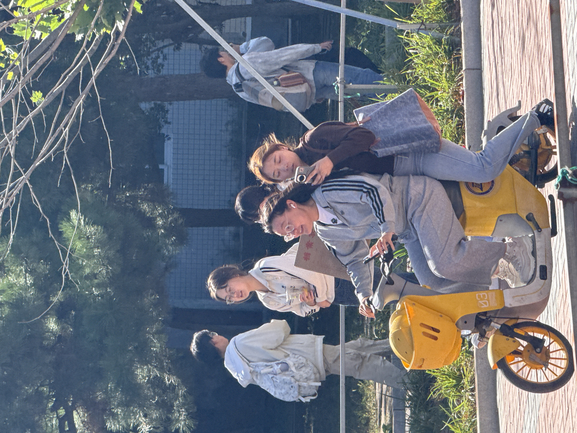 The image captures a sunny outdoor scene, likely in a park or on a street in Guta, China. In the foreground, two young women are seated on a yellow electric scooter. The woman in the driver's seat wears a grey tracksuit and white sneakers, and is holding a mobile phone. The woman behind her wears a brown long-sleeved shirt and grey pants, holding a mobile phone to her ear. A patterned blue tote bag is slung over her shoulder.
Behind them, other individuals are visible. To the left, a young person wearing a white hoodie and cargo pants with a white backpack walks away from the camera. Next to them, a young woman in a white jacket and blue jeans holds a drink and a sign. Further back, another person in a light blue jacket and jeans with a backpack walks from right to left.
The environment features green trees with visible branches, a building with a tiled facade in the background, and a paved walkway. The overall impression is of a casual, everyday moment in a well-lit urban or semi-urban setting.