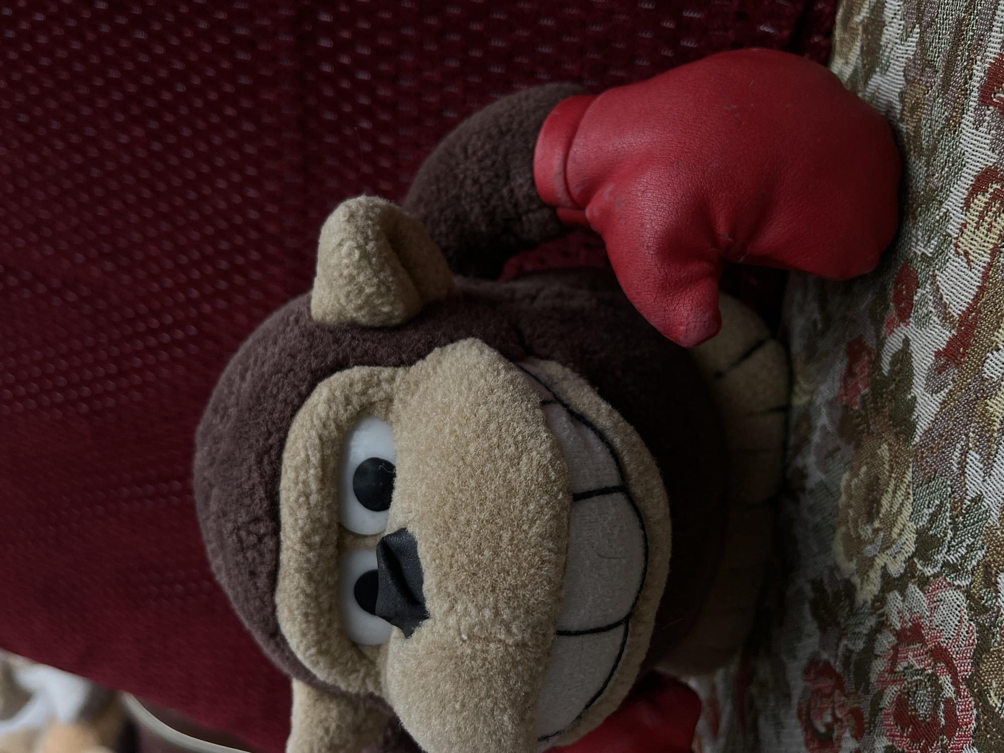 A plush monkey toy with a red boxing glove rests on patterned fabric. The toy has brown fur on its head and body, with a lighter tan color on its face, inner ears, and limbs. It features large, cartoonish eyes and a stitched smile. The red boxing glove is positioned on the monkey's head. The patterned fabric behind the toy is a mix of floral designs in various shades. A red textured background is partially visible to the left of the monkey. The city and location are unknown.