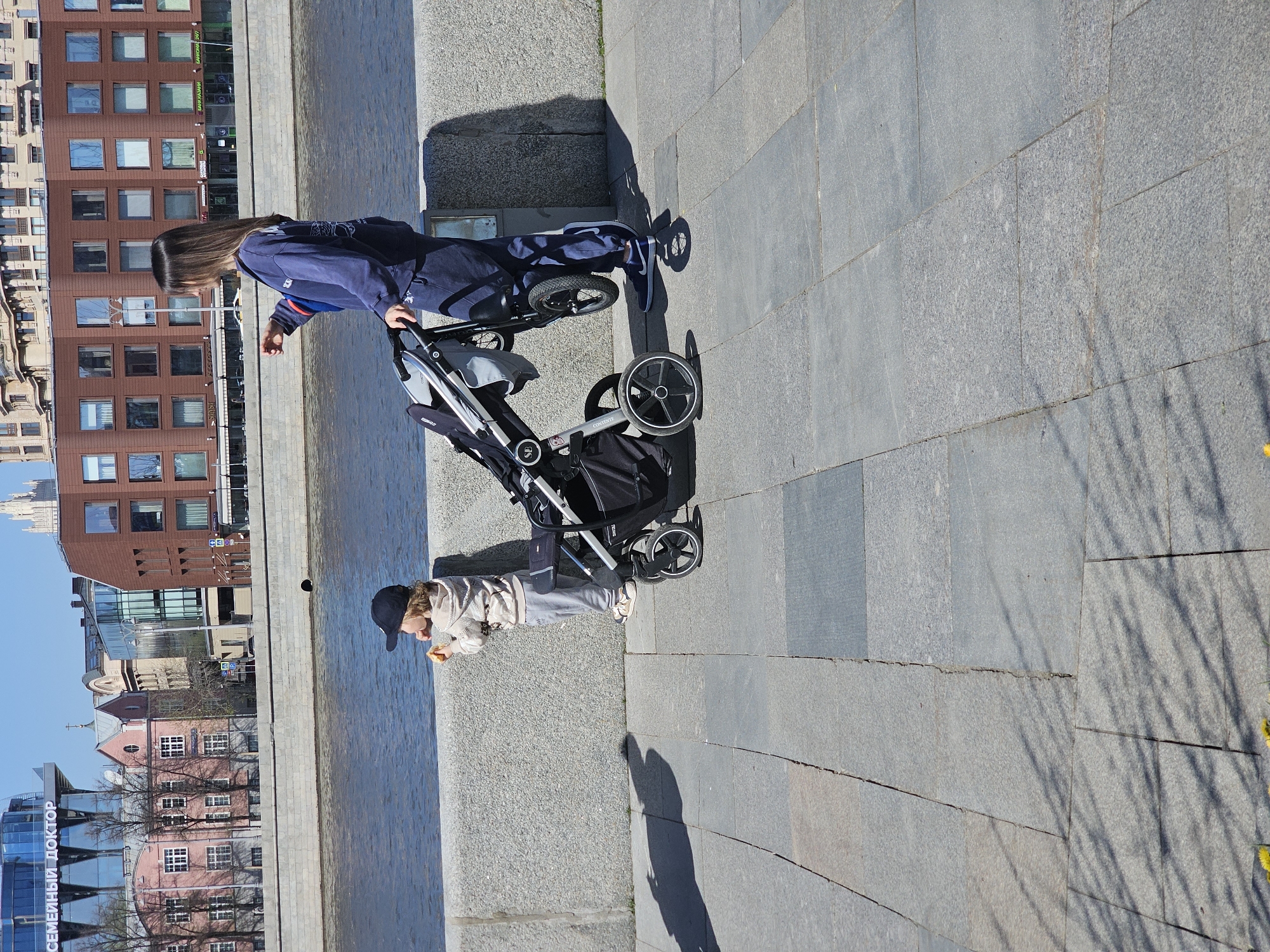 In Moscow, Russia, an outdoor scene depicts a woman pushing a stroller along a paved area next to a body of water. A child stands near the water's edge, eating. Buildings are visible in the background.