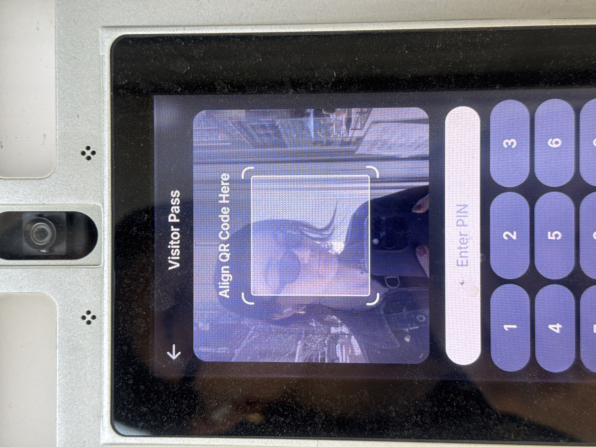 The image shows a close-up of an electronic device with a screen displaying a "Visitor Pass" application. The screen prompts the user to "Align QR Code Here" within a defined square area. The camera appears to be capturing a reflection of a person wearing a dark jacket and possibly glasses within the QR code alignment area. To the right of the screen is a keypad with white and blue buttons for entering a PIN, numbered from 1 to 6. The device is silver in color and appears to be slightly dusty. Based on the application and context clues, it is likely located at an entrance or security checkpoint in a building, potentially in New York City, USA.
