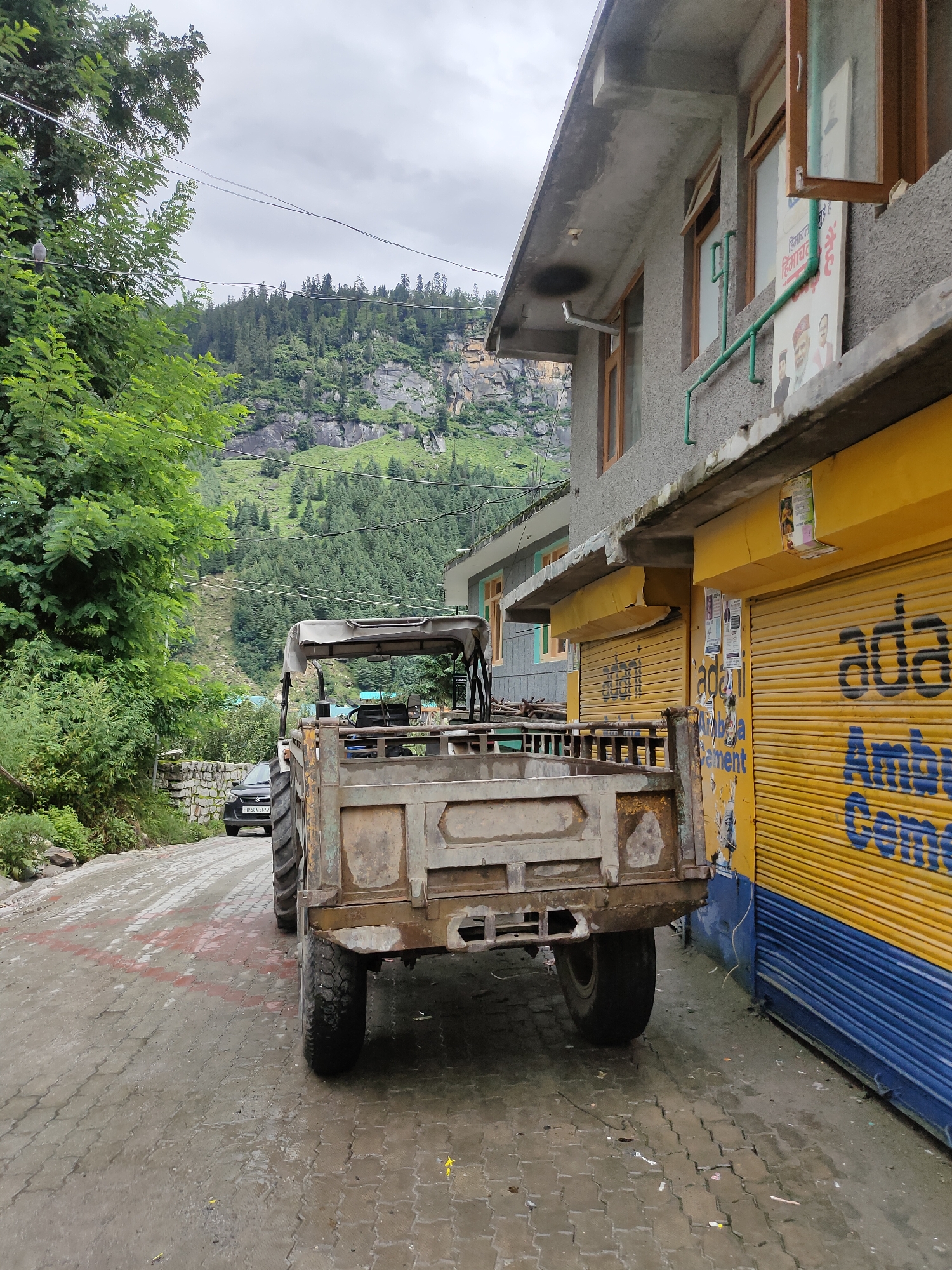 In Burwa, India, a weathered, olive-green tractor with a canvas canopy is parked on a wet, paved road. The tractor's rear is facing the camera, and its large tires are visible. To the right of the tractor is a building with a grey facade and several windows. Below the windows, there are bright yellow roll-up doors with blue accents. The doors are adorned with faded advertisements and branding, including the word "Adani" and "Ambuja Cement" in blue lettering. A green pipe runs vertically down the building's facade. In the background, a dark sedan is visible on the road, and lush green trees line the left side of the path, leading towards a steep, tree-covered mountain under a cloudy sky. The scene appears to be captured on a day with overcast weather, judging by the lighting and the wet road surface.