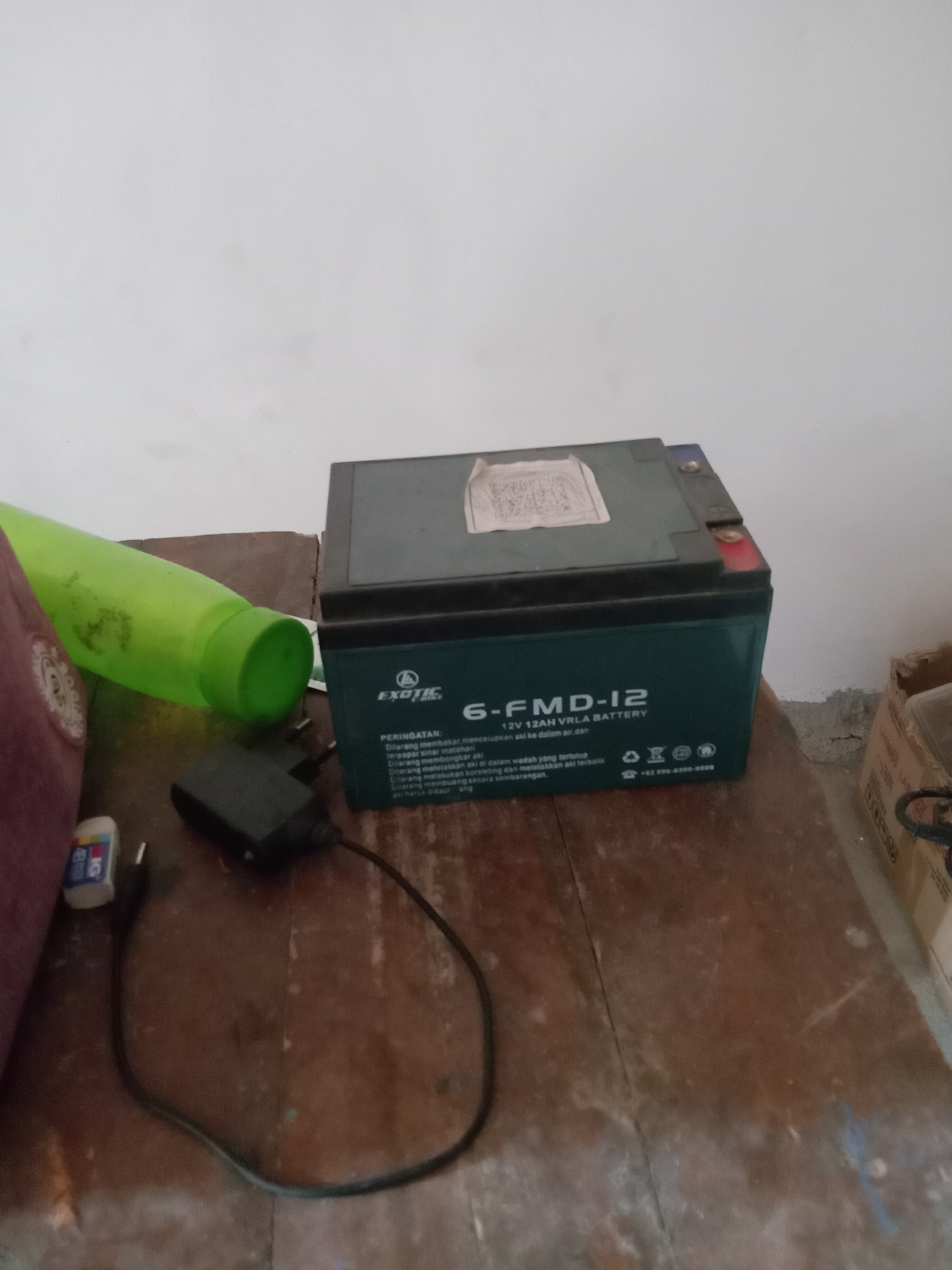 The image shows a dark green, rectangular battery with the label "6-FMD-12 12V 12AH VRLA BATTERY" and the "EXOTIC" logo. Next to the battery is a black power adapter with a coiled black wire. To the left of these items is a bright green plastic bottle, and partially visible on the far left is a maroon object with a patterned texture, possibly fabric. The objects are resting on a wooden surface that appears to be a table or floor, showing signs of wear and cracking. In the background, a white wall is visible, with a portion of a cardboard box on the right side. The scene is illuminated by ambient lighting. There are no people or explicit indications of the city of Pacing, Indonesia, in the image.