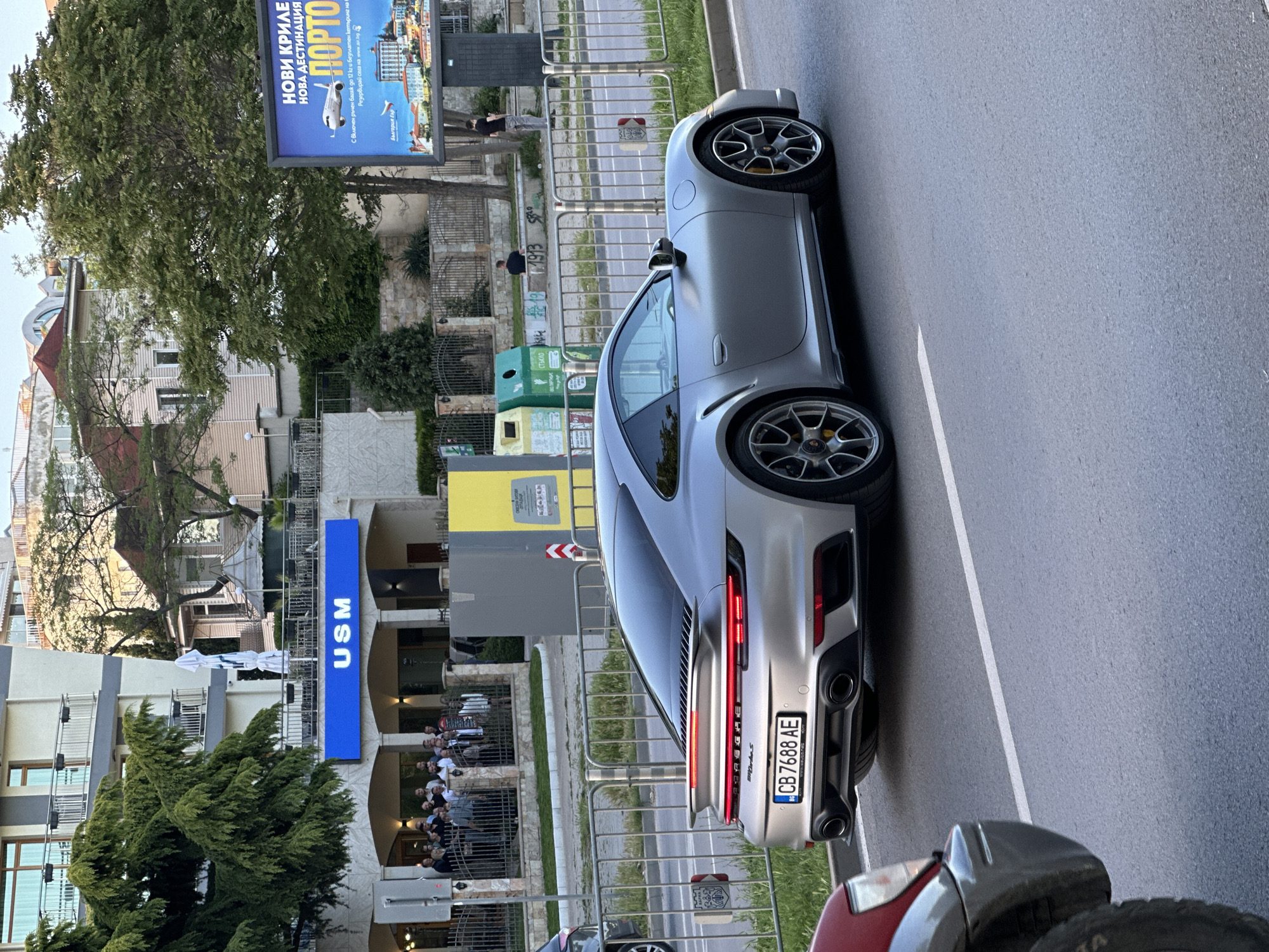 The image shows a silver Porsche 911 Turbo S parked on a street in Varna, Bulgaria. The car has a Bulgarian license plate that reads "CB 7688 AE". Behind the car is a sign that reads "USM", indicating the University of Management and the tall blue advertising signage. To the left, an advertisement for Bulgaria Air promoting flights to Porto, Portugal, is seen. People are congregated near the USM building, suggesting a gathering or event. Trees are visible around the scene, and the street is marked with a white line.