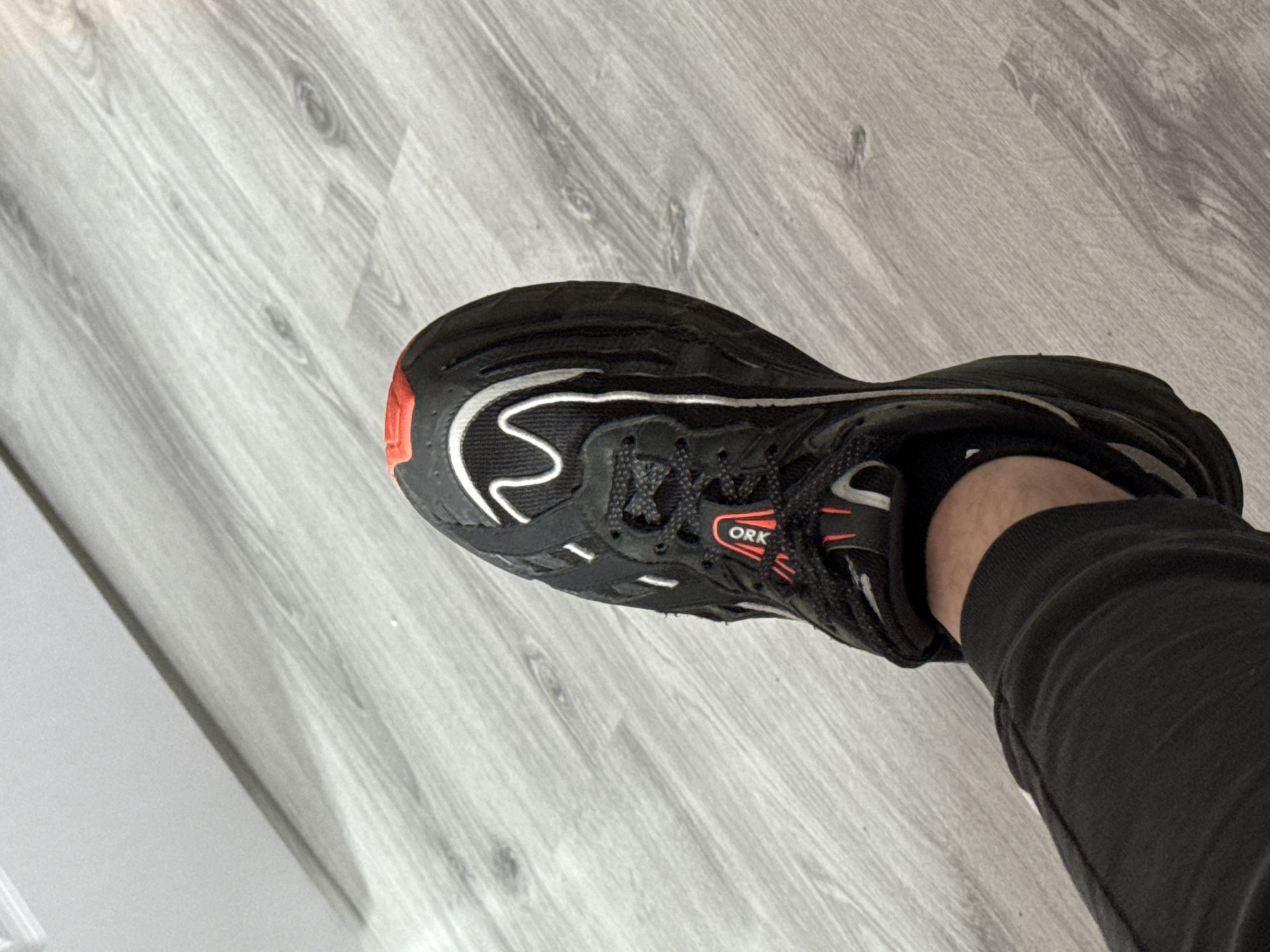 A top-down shot shows a black running shoe on a gray wood-grain floor. A portion of a black sweatpant leg is visible near the ankle. The shoe features white accent lines and a red detail on the heel and near the laces. The inscription "ORK" is visible on the shoe. The environment consists of a grey-toned, wood-patterned floor and a glimpse of a white edge in the lower left corner, possibly a wall. The location or relationship to Stara Zagora, Bulgaria, is not discernible from the visual information.