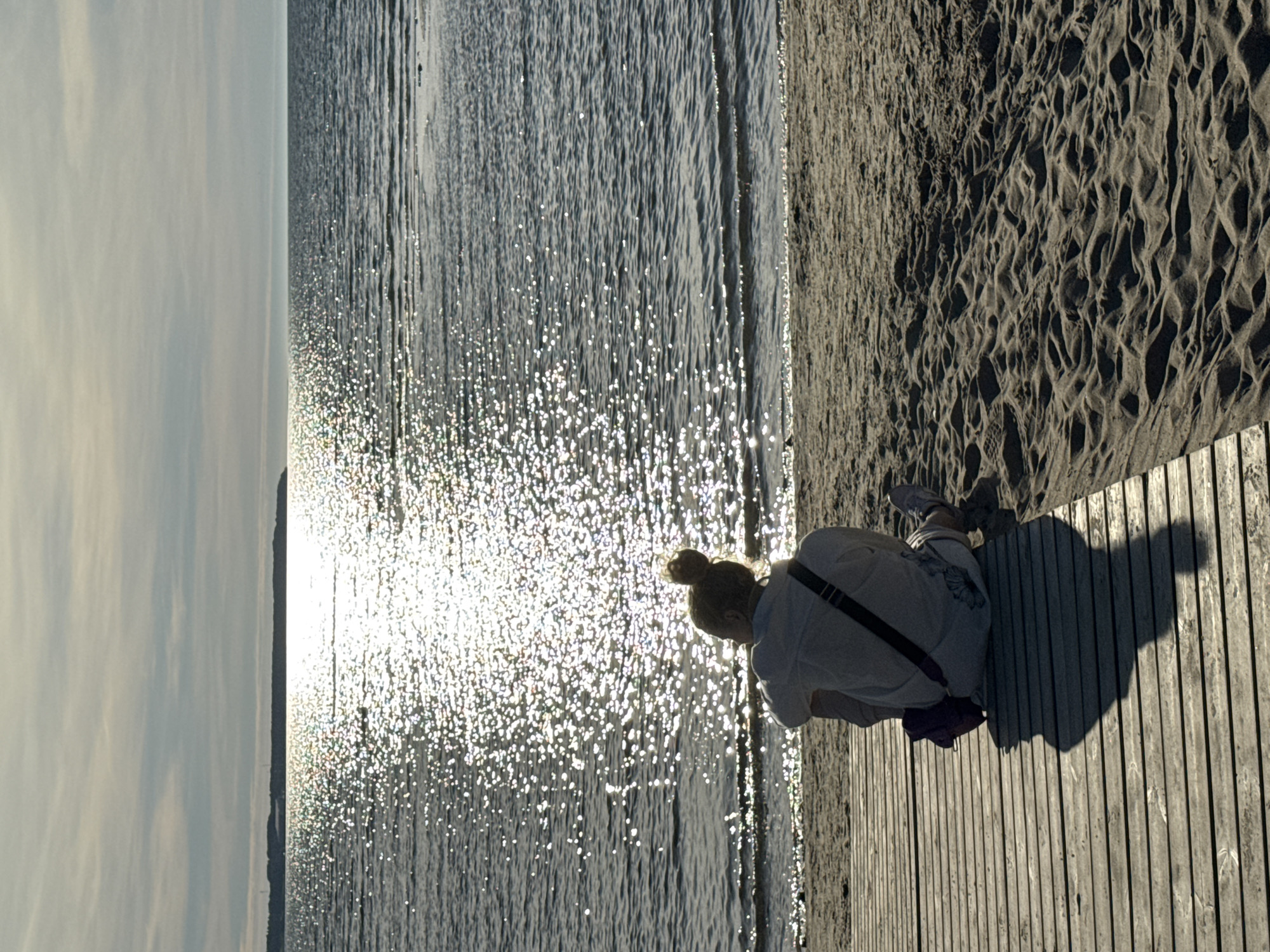 A person is sitting on a wooden walkway on a beach, facing the water. The sun is shining on the water, creating a sparkly effect. The sky is cloudy. There is a landmass visible in the distance. This scene is likely located on the coast of Tallinn, Estonia, potentially near a beach park or a public access point to the Baltic Sea. The person is wearing a light-colored shirt and has their hair in a bun. They have a bag with them. The sand on the beach has ripples and footprints.