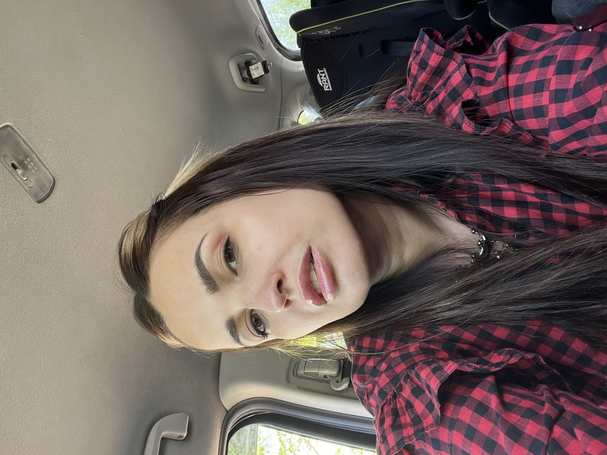 A woman is visible from the upper chest up. She has long, dark hair and is wearing a red and black checkered shirt. The woman is inside a vehicle. The interior of the car is gray, and there is a child car seat in the back. The location is Unknown, Unknown.