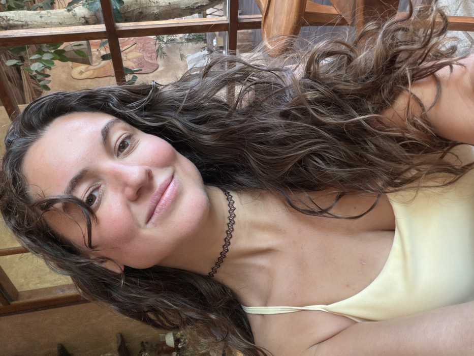 A woman with long, curly brown hair is captured in a selfie. She wears a pale yellow tank top and a black choker necklace. The angle is slightly tilted, emphasizing her face, which has a gentle smile and soft lighting. The background features a wooden frame structure, possibly a window or architectural detail, with glimpses of greenery and foliage suggesting an outdoor or natural setting.