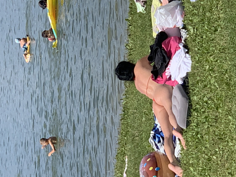 The image captures an outdoor recreational scene on a sunny day at a body of water, described as being in Mykhailivka-Rubezhivka, Ukraine.
In the foreground, an adult female with short, dark hair is lying prone on a patch of green grass facing the water. She is resting on a grey inflatable pillow and is wearing minimal swimwear, specifically a red thong-style bikini bottom and a thin-strapped top, visible across her back. To her left, a large, partially visible brown inflatable beach toy resembling a donut with colorful "sprinkles" rests on the grass. To her right, a pile of various articles of clothing, including pink, black, and white items, along with a white plastic bag, are gathered on a patterned blanket or towel.
In the midground, the water surface shows slight ripples. Three individuals, appearing to be children, are present in the water. On the left, one child is standing chest-deep. Further to the right, another child is lying prone on a yellow inflatable raft, wearing blue swim trunks and a blue swim cap. Adjacent to this raft, a third child is partially visible, also on an inflatable device, with dark hair and possibly a red garment. The scene is brightly lit by sunlight, indicating clear weather.