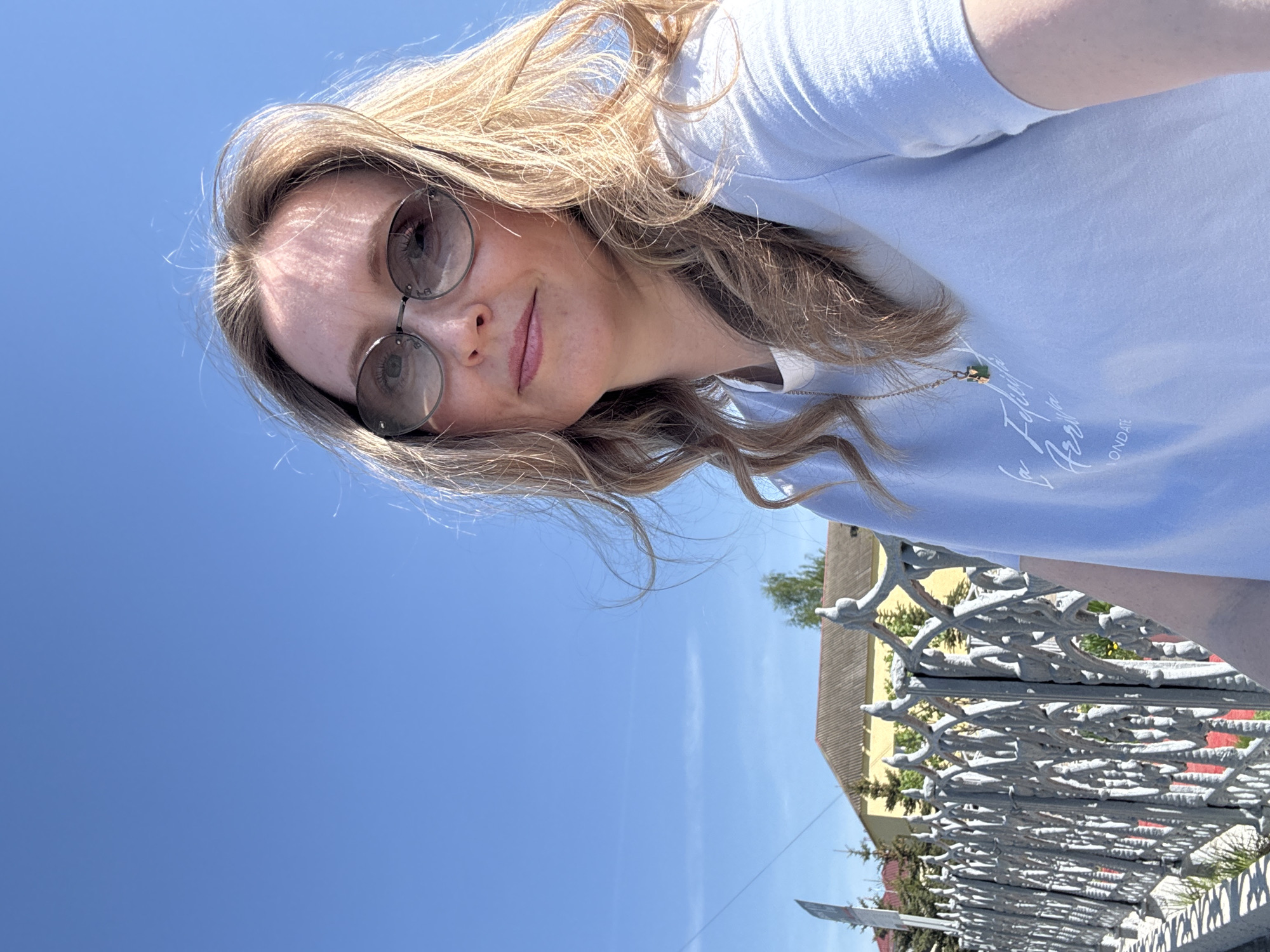 A woman is taking a selfie on a sunny day in Balezino, Russia. She wears a light blue t-shirt, sunglasses, and a gold necklace. Her blonde hair is loose. Behind her is a light-colored building and a decorative grey metal fence. The sky is clear blue.