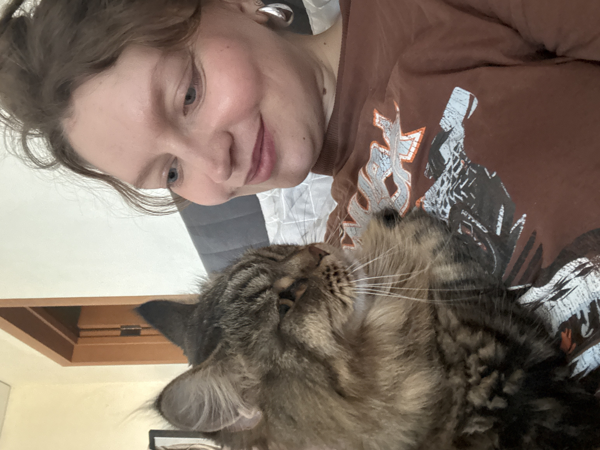 A woman with fair skin, light blue eyes, and light brown hair is looking down at a tabby cat nestled against her chest. The woman is wearing a brown t-shirt with a graphic design on it and silver hoop earrings. The cat's eyes are closed, and its whiskers are prominent. The background includes a glimpse of a white and grey patterned surface and a wooden window frame. The location of Izhevsk, Russia, is not ascertainable from the image.