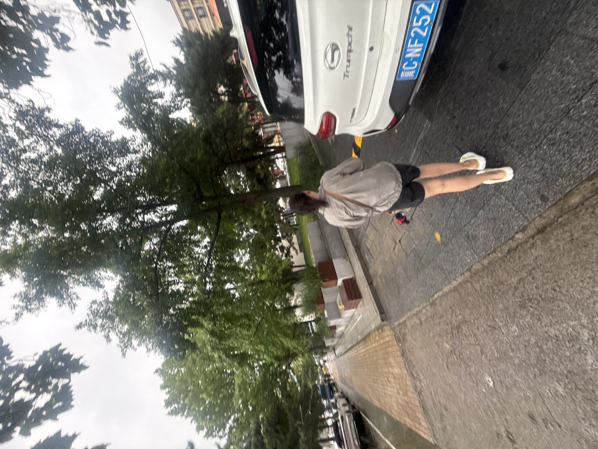 A person walks on a street in Qinhuangdao, China, with a white SUV parked on the right side of the road. The person, seen from behind, is wearing a light-colored jacket and dark shorts, with white shoes. The SUV has a license plate that reads "C-NF 252". The street is lined with trees and a sidewalk. The weather appears overcast.