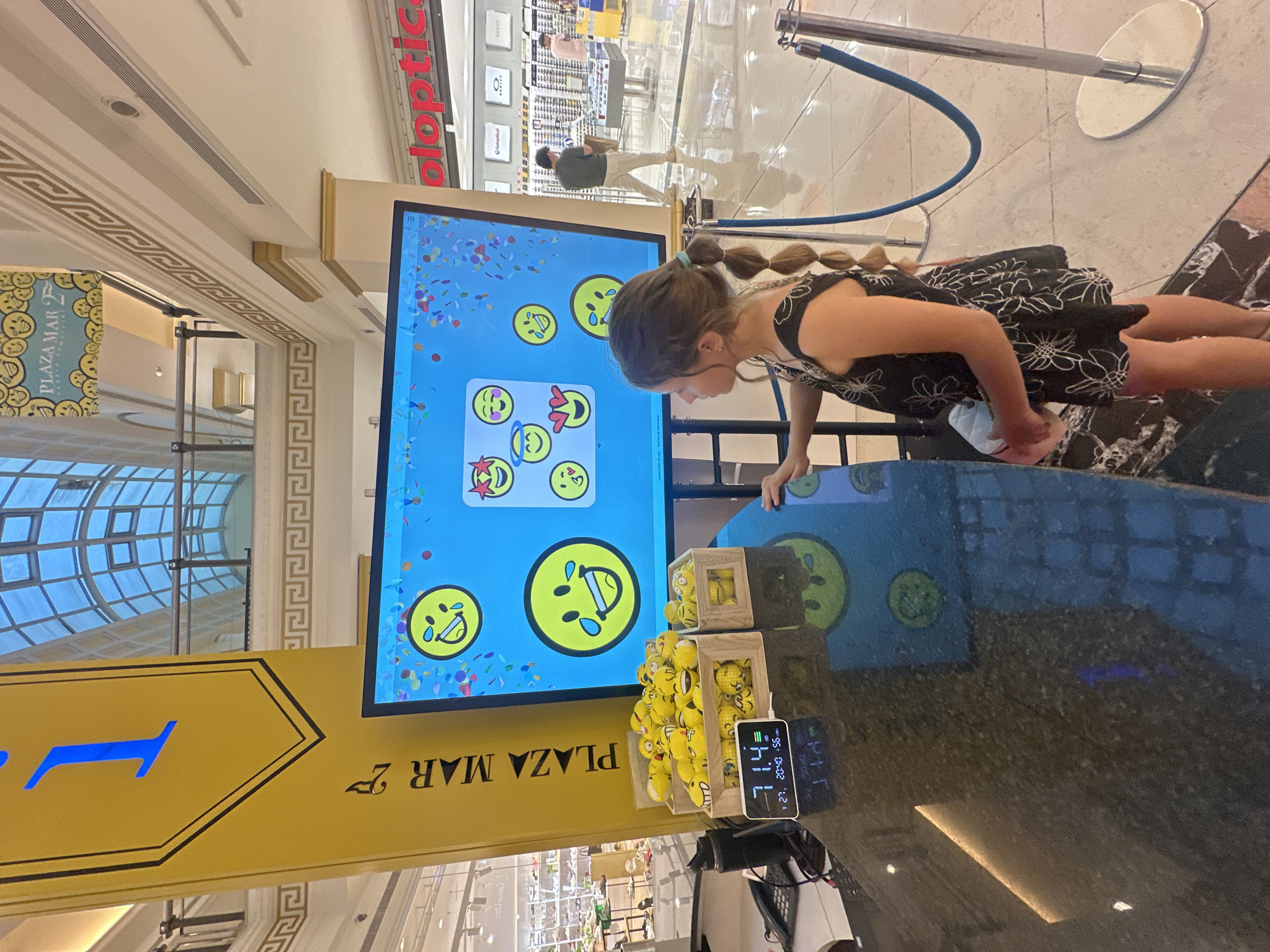 A young girl, seen from a side-rear angle, is focused on a large vertical digital screen in what appears to be a brightly lit shopping mall. The image is oriented vertically (rotated 90 degrees clockwise). The girl has braided hair and wears a dark dress with white floral patterns. She leans forward, her hands near the base of the screen, suggesting interaction with the display.
The screen displays a game or interactive presentation featuring various emoji faces, with confetti scattered across the background. To the right of the screen, two wooden boxes are filled with numerous small yellow balls, each featuring an emoji face. A small digital device rests on the counter next to the boxes, displaying "71.4", "27", "20:00" (suggesting 8:00 PM), "55%", and "31.1".
The setting is an indoor commercial space, characterized by a contemporary design. Architectural details include ornate gold Greek key patterns on ceiling or wall trim and a large glass-paned ceiling or window structure allowing natural light to enter. In the background, metal stanchions with a blue rope are visible, likely forming a queue line. Other shoppers can be seen in the distance, out of focus.
Visible text includes "PLAZA MAR 2" and "PLAZA MAR" on yellow signs, indicating the name of the shopping center. The red text "oloptic" and blurry "GUCCI" and "O Soleoptica" signs are also visible in the background, likely store names. The overall atmosphere suggests an engaging and playful environment within a mall.