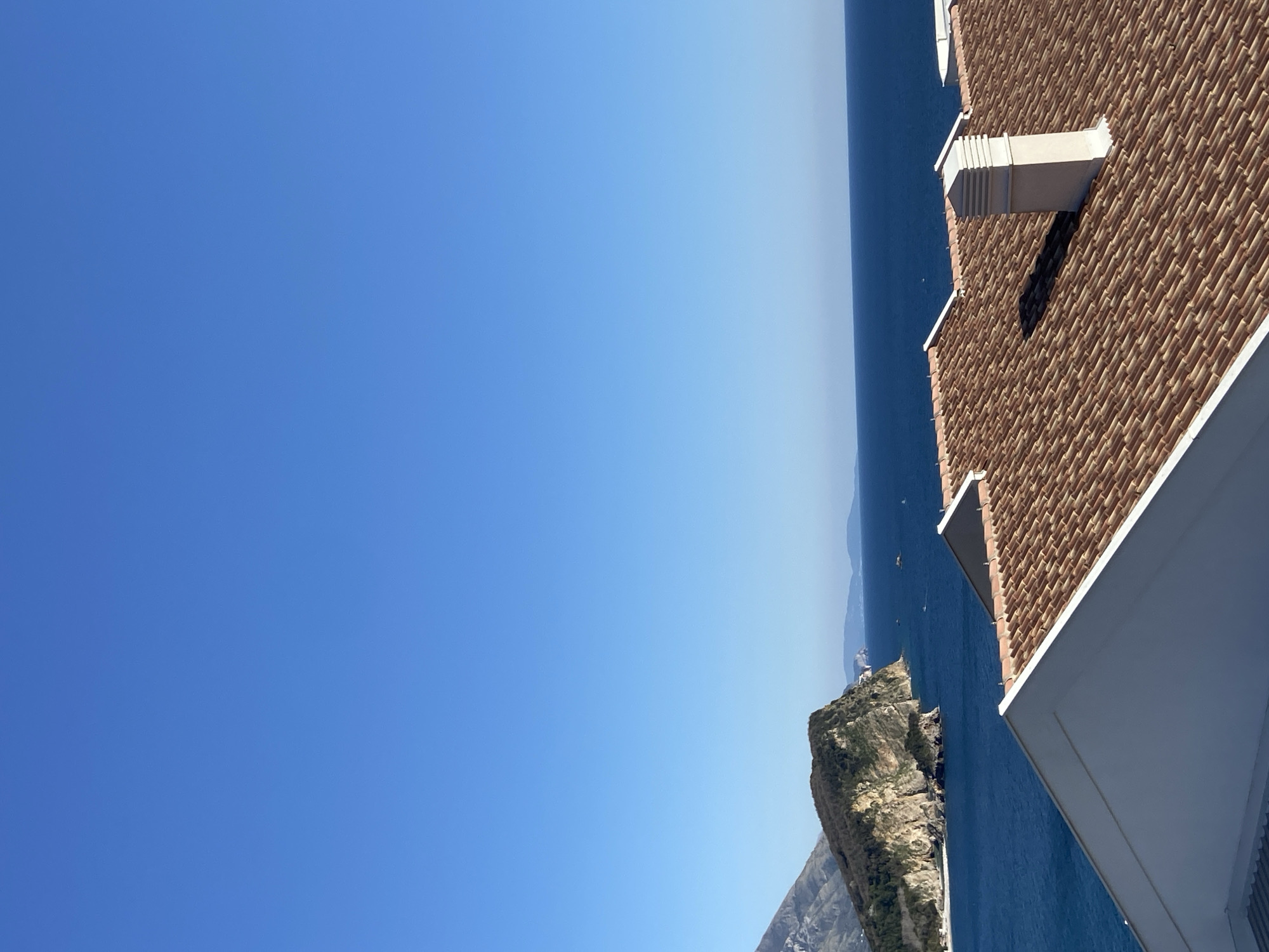 Here is a description of the image:
The photograph shows a landscape view, including a building structure, the sea, cliffs, and sky. The scene appears to be near the coast. The sky is clear and blue, transitioning to a slightly lighter shade near the horizon where it meets the sea. The sea is a deep blue color, with a few small boats visible. Adjacent to the sea is a cliff with green vegetation on its surface. The right side of the image includes a portion of a building with a brick-tiled roof and white architectural elements, suggesting it could be part of a coastal villa or a hotel. The image's orientation is rotated, which offers an unconventional perspective. The precise location in Budva, Montenegro, is difficult to determine without additional landmarks.