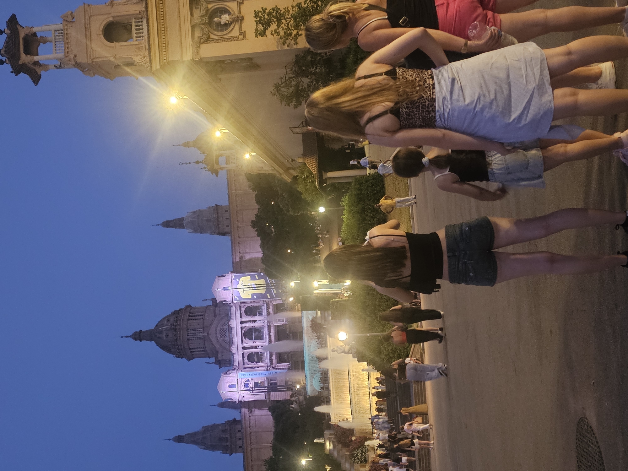 This image depicts the Magic Fountain of Montjuïc in Barcelona, Spain, at dusk. A group of people, including several women and a young girl, are standing in the foreground facing a large building with domes and towers, illuminated by purple and yellow lights. In the middle ground, the Magic Fountain is active, with water cascading down multiple tiers, and many people are visible on the steps and surrounding areas. The sky is a deep blue, transitioning from daylight to night. The architecture suggests a grand, public space, likely a museum or palace, as seen in the background.