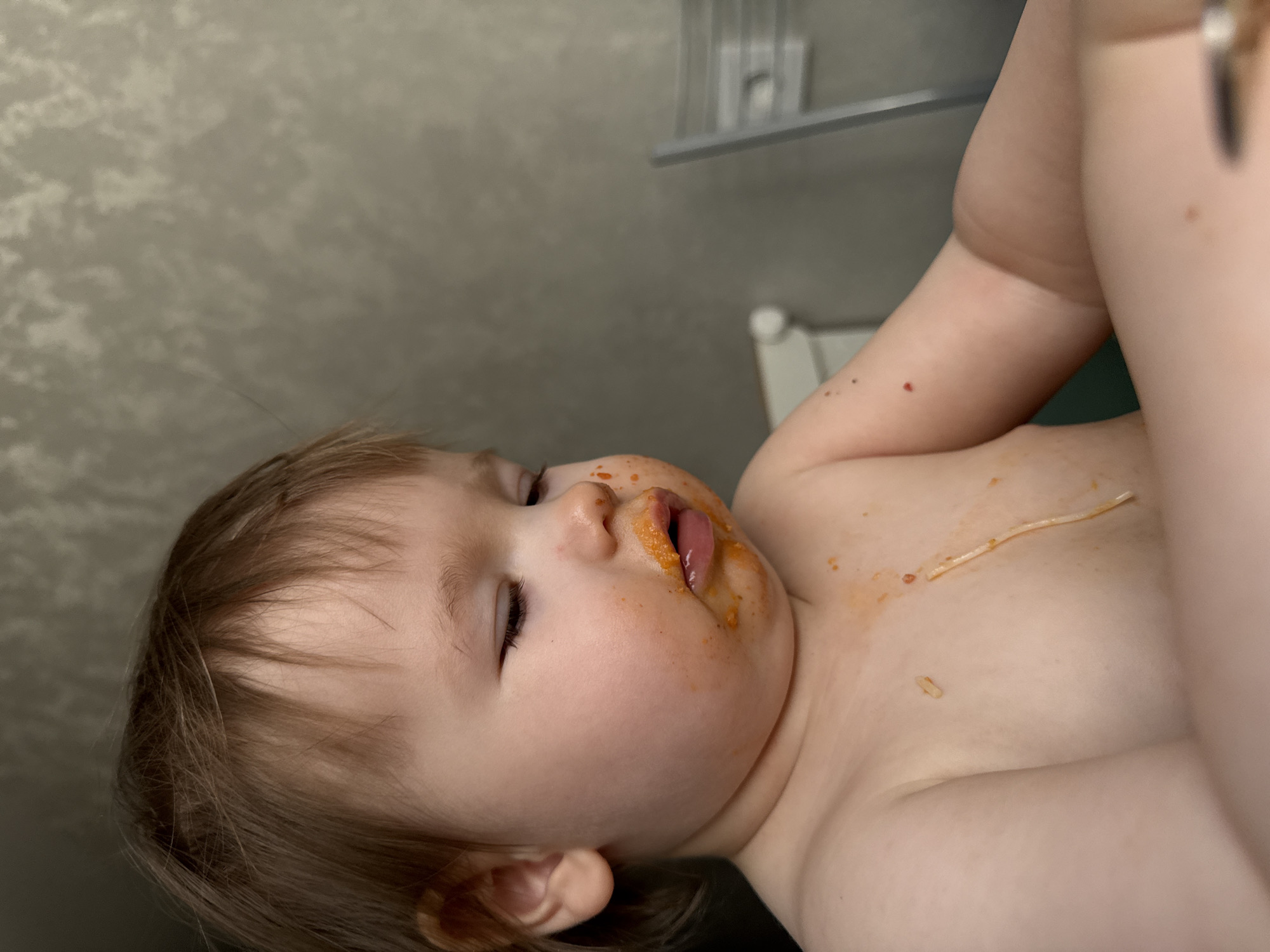 A young child is depicted, lying on their back, with their body oriented towards the lower right of the frame. The child is covered in what appears to be tomato sauce, particularly around the mouth and chest. A single strand of spaghetti is visible on the child's chest. The child's eyes are partially closed. In the background, there is a textured, grey-toned wall and a chrome towel rack. The location or city is not discernible from the image.