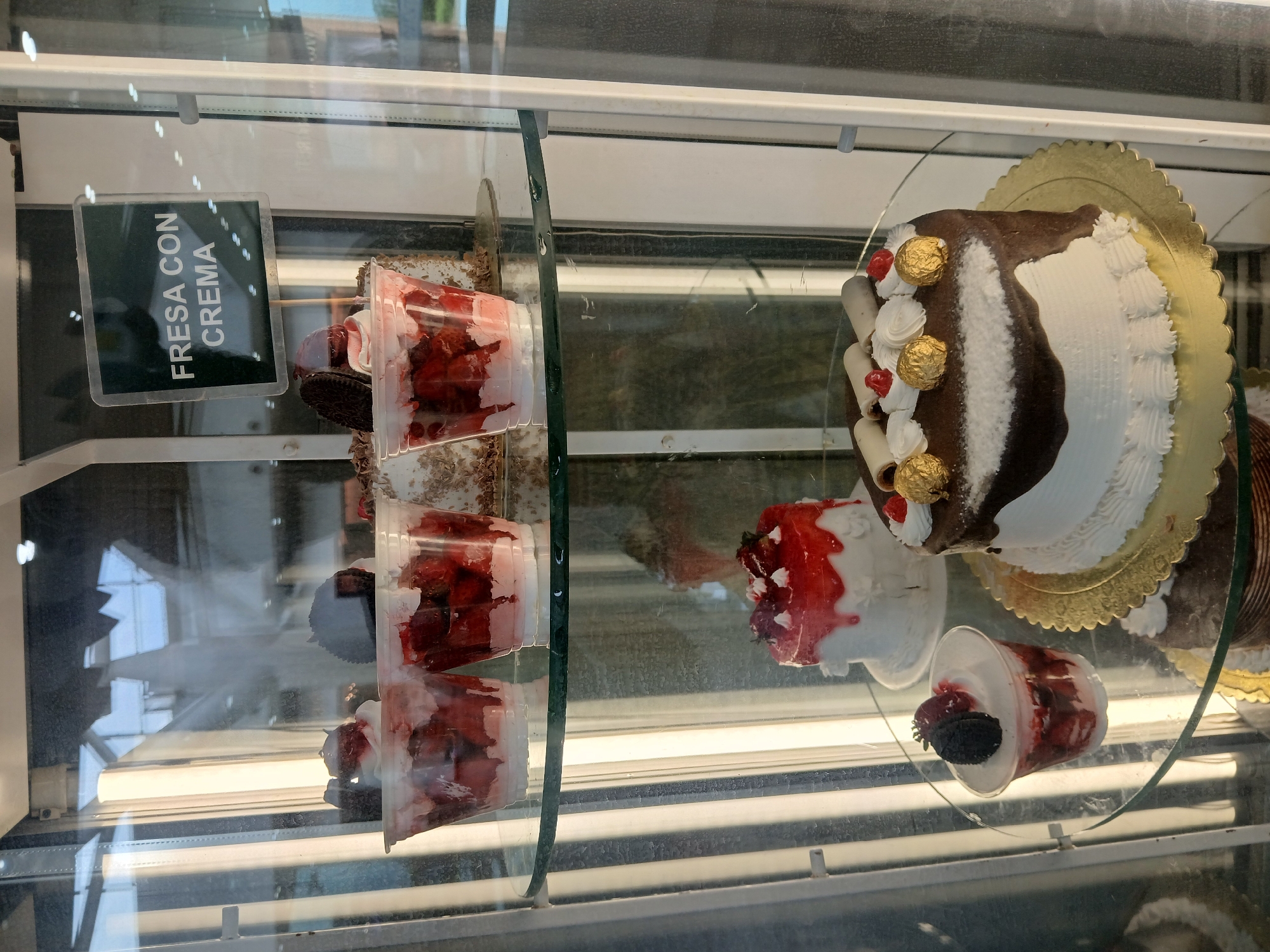 A display case contains several desserts, including a cake with chocolate ganache and white frosting, adorned with Ferrero Rocher candies and rolled wafers. Smaller containers hold strawberry and cream parfaits, one of which is labeled "Fresa Con Crema." The scene is located in Ciudad Guayana, Venezuela, based on the Spanish-language signage. The display case has glass shelves and is likely located in a bakery or dessert shop.