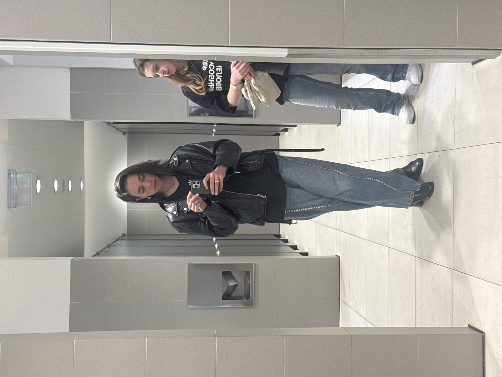 The image shows a person in a public restroom taking a mirror selfie. The person is wearing a black leather jacket, a black shirt, and blue jeans with embellishments. They are holding a green smartphone. The restroom has gray tiles, white walls at the top, and metal fixtures, including a hand dryer. The image is taken in front of a mirror, and another person is visible in the reflection, wearing a dark top and jeans, holding a light-colored bag. The scene is indoors. The location is not discernible as Belgrade, Serbia, based on visual information alone.