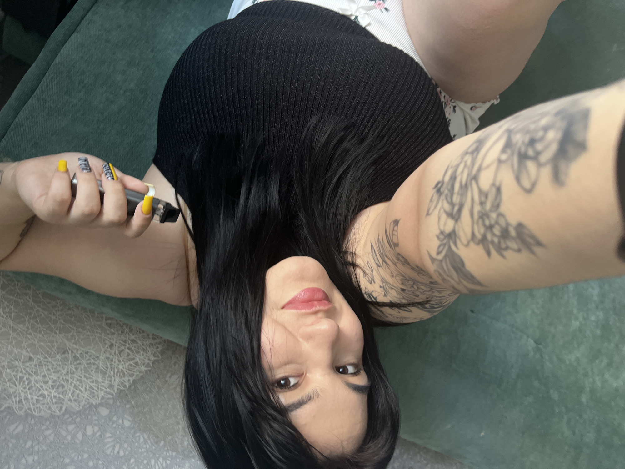 A woman lies on a green couch holding a vape pen with yellow-colored nails. She has black hair and tattoos on her right arm. The location is not discernible from the image.