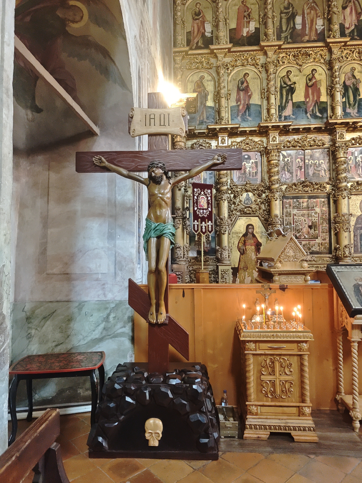 The image depicts an interior view, likely of a church or religious building, in Chelganovo, Russia. A large wooden crucifix with a figure of Christ is prominently displayed on the left, positioned atop a black, rock-like base with a skull on the front. Behind the crucifix is a painted wall with indistinct figures. To the right is a wooden cabinet adorned with a cross, featuring numerous lit candles on top. Behind the cabinet is a highly decorated golden iconostasis filled with religious paintings. To the left of the cross is a small wooden table. The floor is tiled with brown tiles. The overall scene suggests a religious or ceremonial setting.