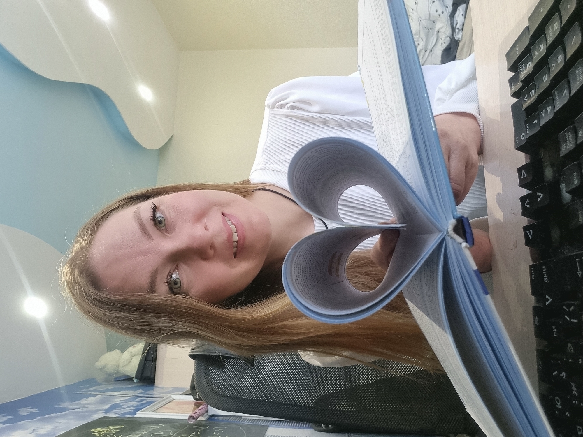This image is a close-up, low-angle selfie of a young woman, captured indoors and rotated 90 degrees clockwise. The woman, with long, light brown hair, striking green eyes, and a radiant smile, looks directly at the camera. She is wearing a white ribbed long-sleeved top. Her left hand is prominently featured, holding an open light blue book or binder whose pages are artfully bent to form a heart shape, a central focal point of the image.
In the immediate foreground, to the right, a black computer keyboard with visible Cyrillic characters (such as "ы", "ф", "ц", "у", "к", "е", "н", "г", "ш", "з", "х", "ъ", and Latin "V") rests on a light-colored desk. A dark, mesh-textured object, possibly a part of a chair or bag, is visible in the lower-left corner. On a blue-patterned desk surface, below the woman, lie papers and a small pink cylindrical object.
The background features a light blue wall on the left, adorned with a modern, wavy white ceiling fixture containing integrated lights. A light yellow wall is visible on the right. Behind the woman's shoulder, a blurry fabric with a pattern resembling bears can be seen. The scene suggests an indoor setting, likely a workspace or study area, in Chaikovsky, Russia. The woman's expression conveys happiness and engagement.