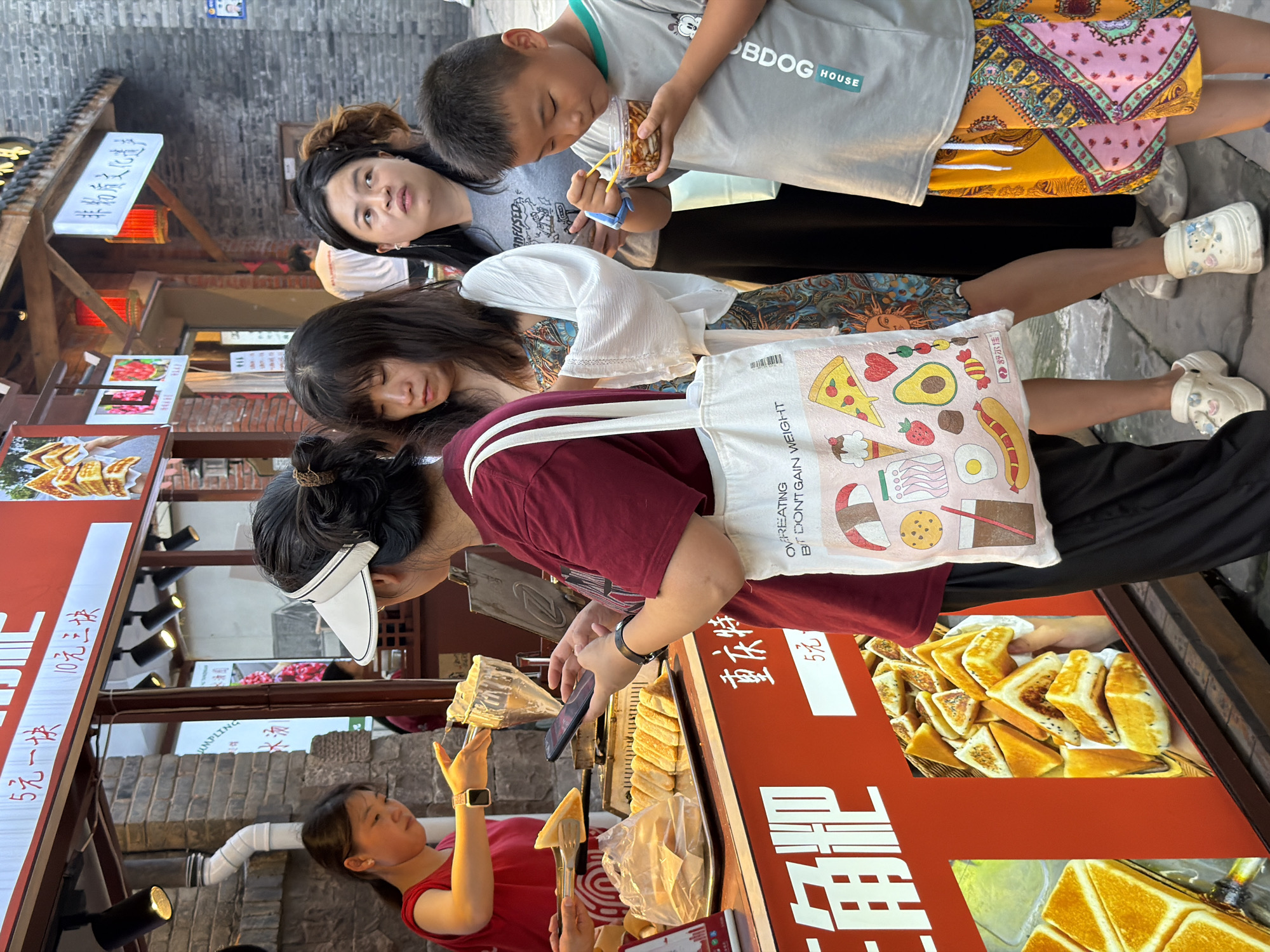 In the Yuzhong District of China, a street vendor sells a sweet dish resembling golden fried bread. The vendor, a woman in a red shirt, prepares the food with tongs while a customer, a woman wearing a maroon shirt and a white visor, looks at her phone. Behind them, several other people stand, including a young boy in a grey t-shirt and colorful shorts, who is eating from a small container. The stall displays images of the food and price information in Chinese characters. The environment appears to be a bustling marketplace, with brick buildings and outdoor lighting.