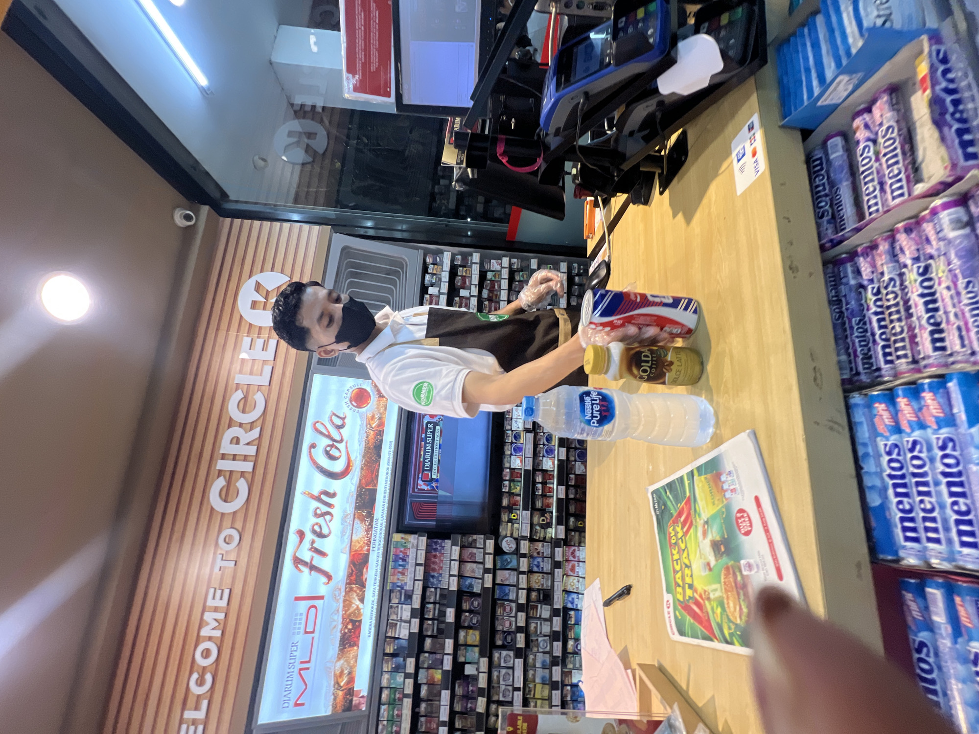 A male cashier, wearing a white polo shirt, a black face mask, and a brown apron, stands behind a wooden counter at a Circle K convenience store. He is wearing clear plastic gloves. In front of him are a bottle of Nestlé Pure Life water, a can of drink, and a bottle of Golda coffee. A promotional flyer for Circle K is also visible on the counter. Behind the counter, shelves are stocked with various products, including cigarettes. A computer monitor and a credit card reader are also present. The store signage, "WELCOME TO CIRCLE K," is visible above the counter. The scene appears to be in Denpasar, Indonesia, based on the context provided by the user. The overall environment is a retail setting with fluorescent lighting.