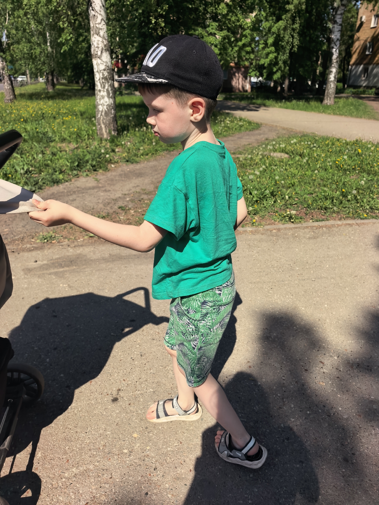 A young boy stands on a concrete path, pushing a stroller. He is wearing a green t-shirt, patterned green shorts, and sandals. A black baseball cap sits atop his head. Sunlight casts shadows on the path. The background shows a grassy area with trees and a building in the distance. The location is Sterlitamak, Russia.